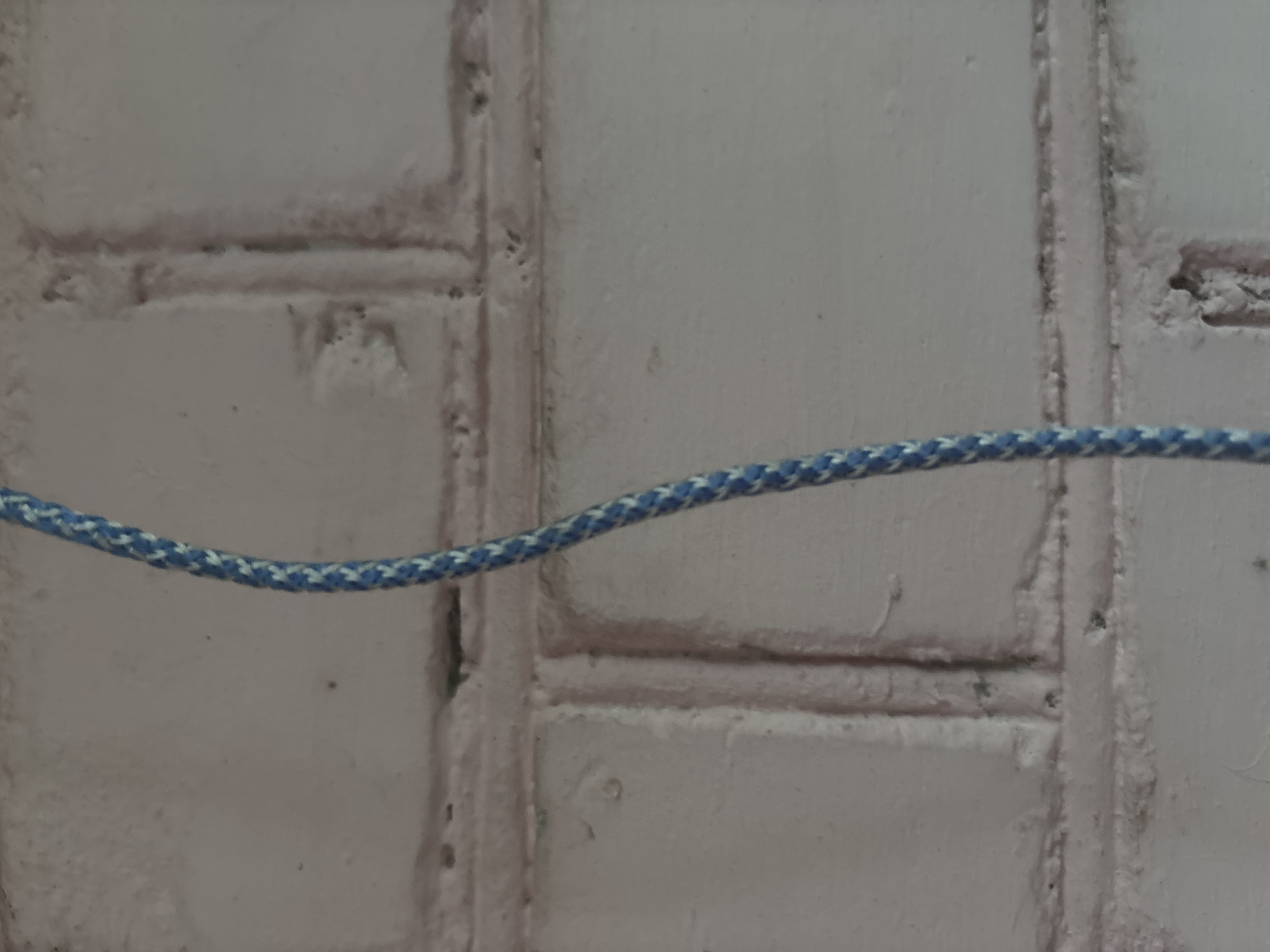 The image shows a close-up of a blue and white braided rope hanging vertically against a white brick wall. The rope is slightly curved. The brick wall appears to be rough and textured, with visible mortar lines. The lighting suggests an indoor or shaded outdoor environment. The scene is devoid of people or any discernible activity. The location is unknown.