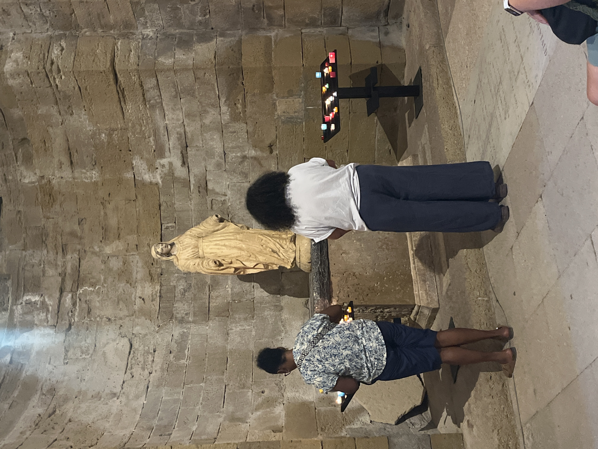 Two individuals stand in what appears to be a stone church interior. A statue of a robed figure, likely religious, stands prominently against a stone wall. In the foreground, two black racks hold numerous small, lit candles, some in glass holders. One person, viewed from the back, wears a patterned shirt and shorts. The second person, also viewed from the back, wears a white t-shirt and wide-legged pants. The overall environment suggests a place of worship. This scene is located within the former cathedral of Saint-Pierre-et-Saint-Paul, now known as the Cathédrale de Villeneuve-lès-Maguelone, in Villeneuve-lès-Maguelone, France, which is partly ruined but still retains elements of its former grandeur.