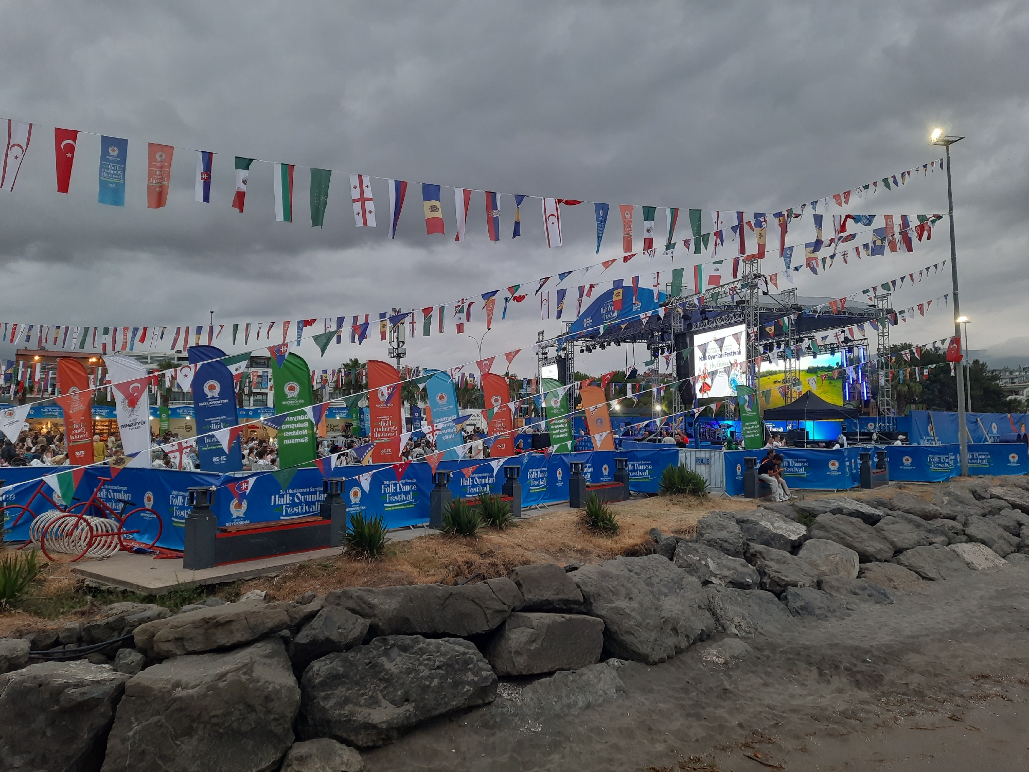 This image depicts an outdoor festival scene in Atakum, Turkey, under a cloudy sky. A stage with lighting and screens is visible in the background, along with numerous flags of various nations strung across the area, suggesting an international event. Several blue banners with text that appears to read "Folk Dance Festival" are prominently displayed. In the foreground, large rocks line a shoreline, with a red bicycle parked on a concrete area. People are scattered throughout the scene, some seated in front of the stage and others milling about. The overall atmosphere is that of a community gathering or celebration.