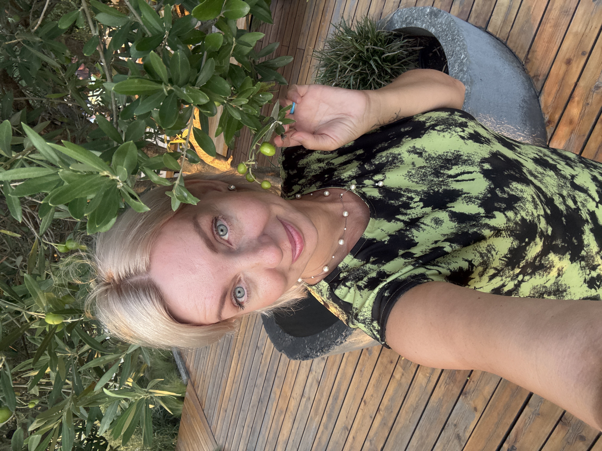 A blonde woman with blue eyes is pictured from the chest up, holding an olive branch with green olives. She is wearing a black and lime green tie-dye shirt and a pearl necklace. She is standing on a wooden deck next to a large concrete planter with a plant inside. Behind her is an olive tree with more green olives and leaves. The scene appears to be outdoors in the city of Sirius, Russia.