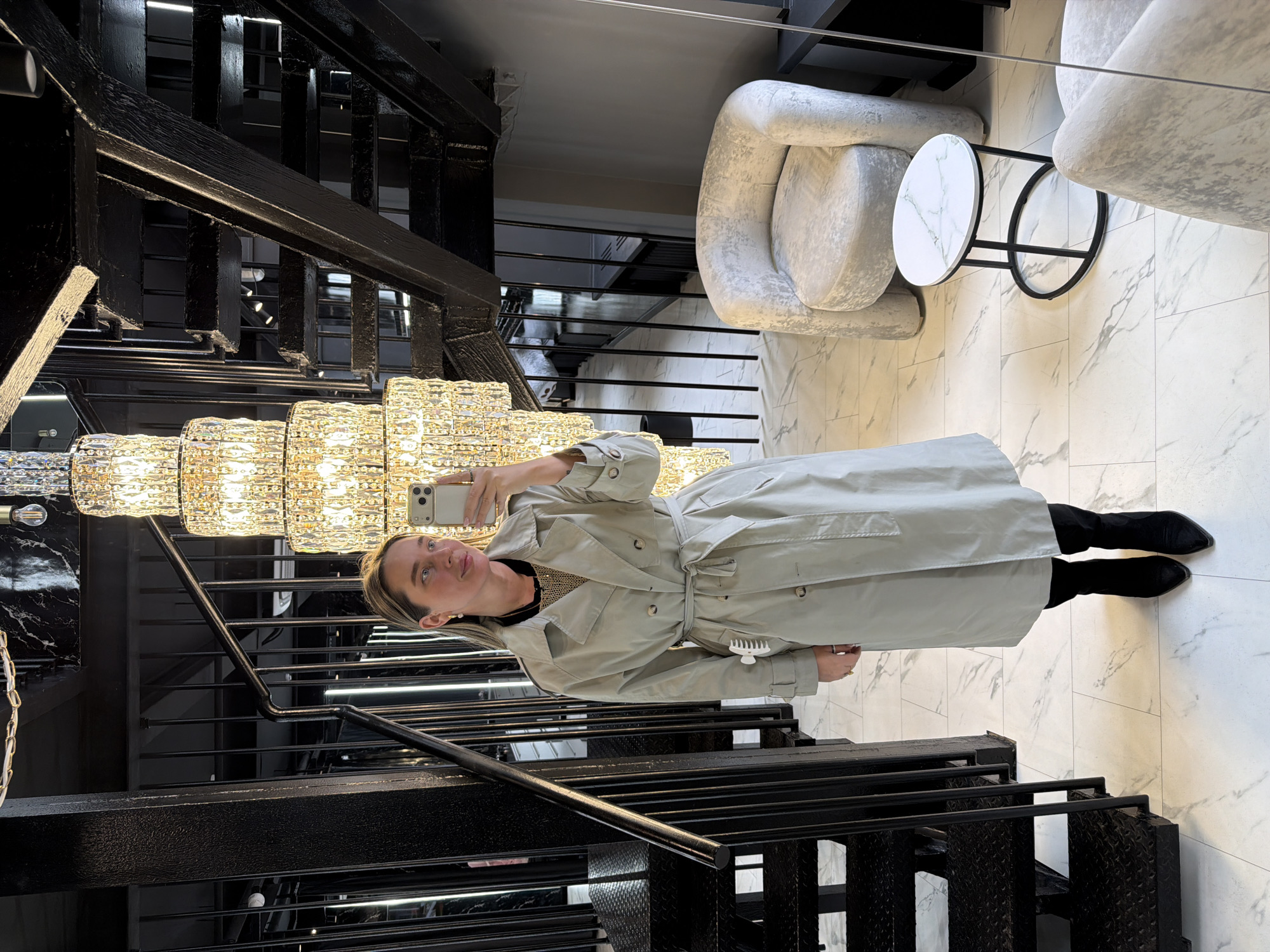The image shows a woman taking a selfie. She is standing on a marble floor, wearing a light grey trench coat and black boots. She is standing in front of a black metal staircase. A large chandelier hangs from the ceiling above her. In the background, there are two light grey armchairs and a white marble-topped side table. The location is Moscow, Russia.