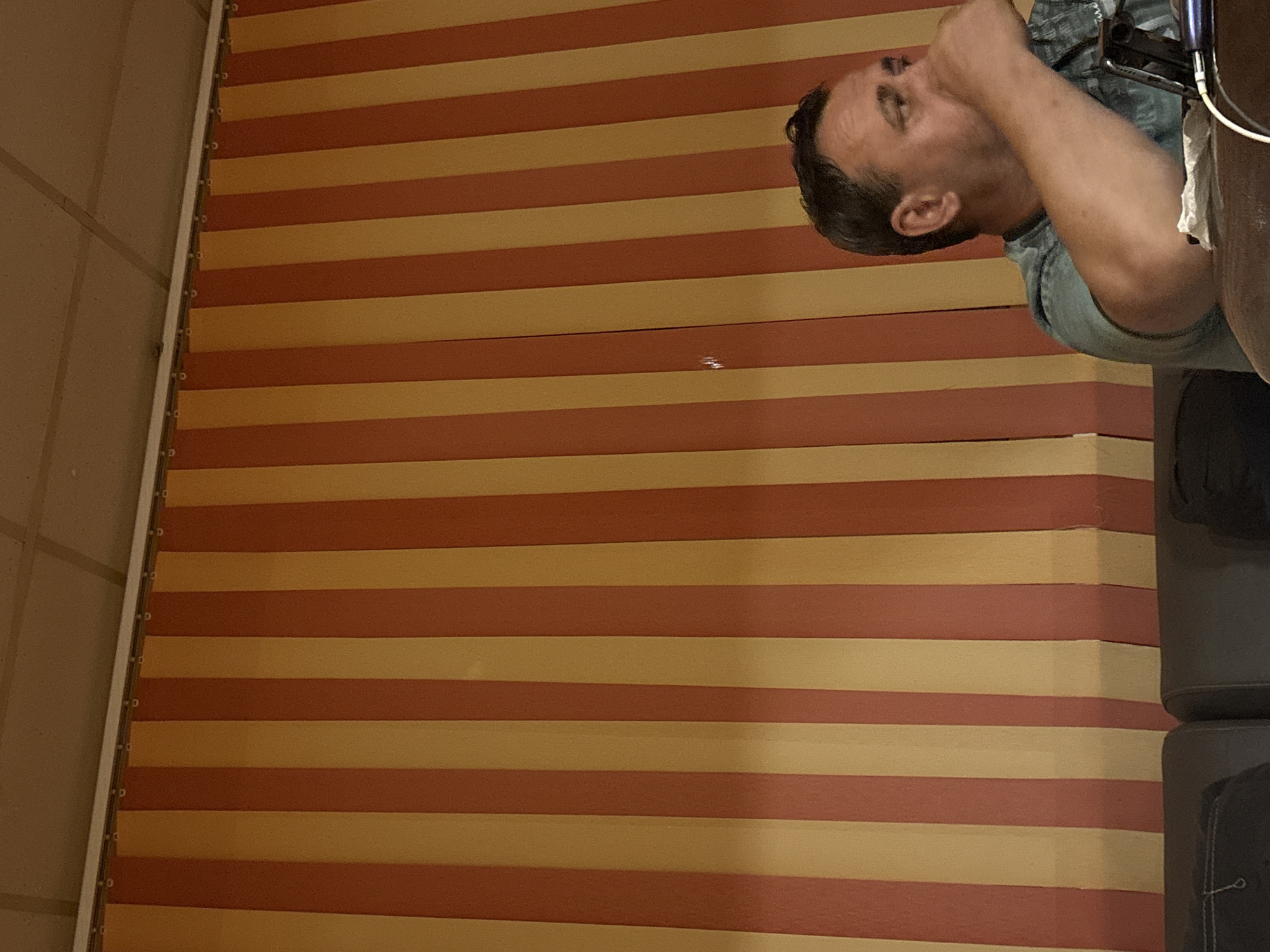 This media file is a portrait of a man in Tervel, Bulgaria. The man appears to be of adult age and is wearing a blue t-shirt. He is positioned in the upper right quadrant of the image, with his head tilted back and his hand touching his face. The background consists of horizontal stripes in alternating shades of orange and yellow. To the right of the man, there are dark gray objects, possibly furniture, with white paper tucked into one of them. The lighting suggests an indoor setting, possibly during the daytime. The man's expression is not clearly discernible due to the angle and his hand partially obscuring his face. There is no visible text in the image.