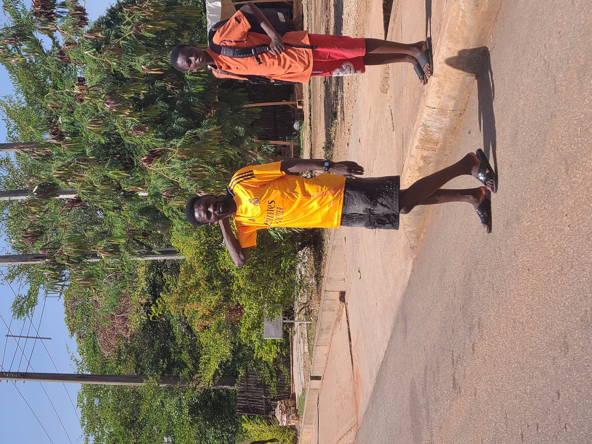 Two young men are standing on the side of a paved road. One wears a yellow shirt and black shorts, while the other has an orange shirt, red shorts, and a backpack. Both wear sandals. They are near a sidewalk curb, with lush green vegetation and utility poles visible in the background. The location is suggested to be Jos, Nigeria, but that cannot be verified solely from the visual content of the image.