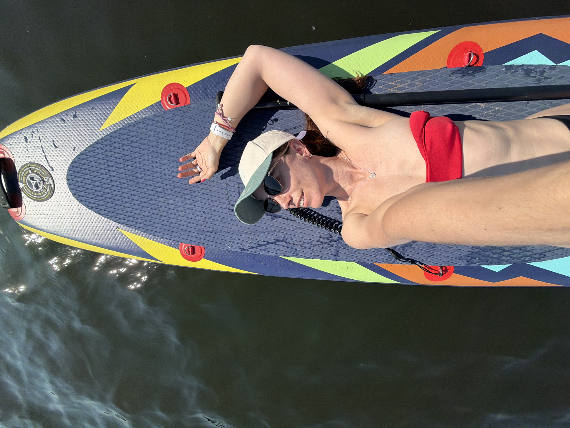 A woman is lying on a paddleboard in the water. She is wearing a red bikini top, a hat, and sunglasses. She has a watch and bracelets on her left wrist. The paddleboard has a blue textured surface with yellow, orange, and green accents. A paddle lies next to her on the board. The water is dark and reflects sunlight. The location is not Birobidzhan, Russia.