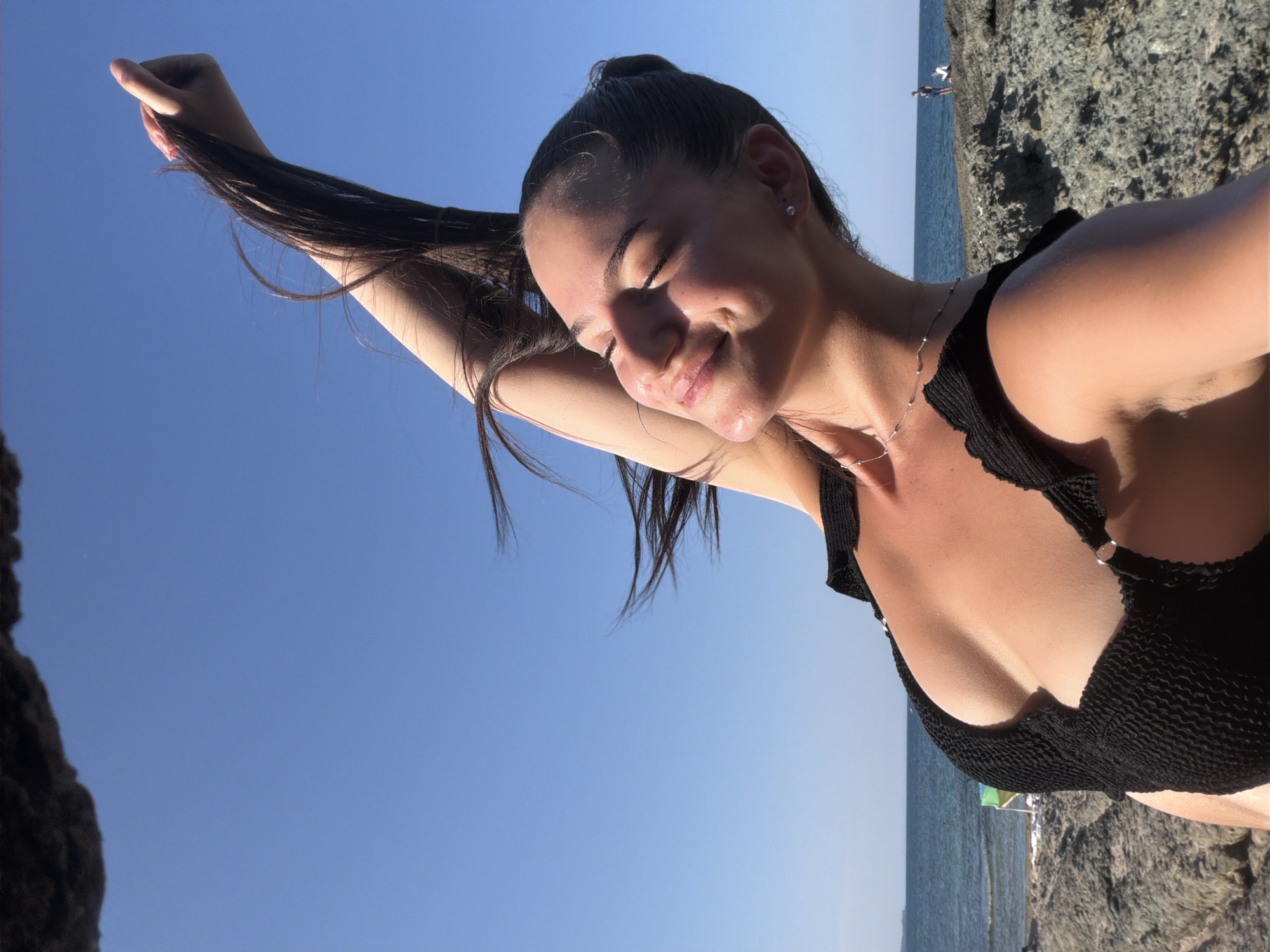 A young woman with dark hair, wearing a black bikini, is captured in a low-angle shot outdoors. Her eyes are closed, and she has a slight smile on her face, her head tilted back with her arm raised, holding a lock of her hair. The background shows a clear blue sky, the sea, and a rocky coastline. The scene appears to be a beach in Lozenets, Bulgaria, given the characteristic rocky shoreline and the presence of the Black Sea.