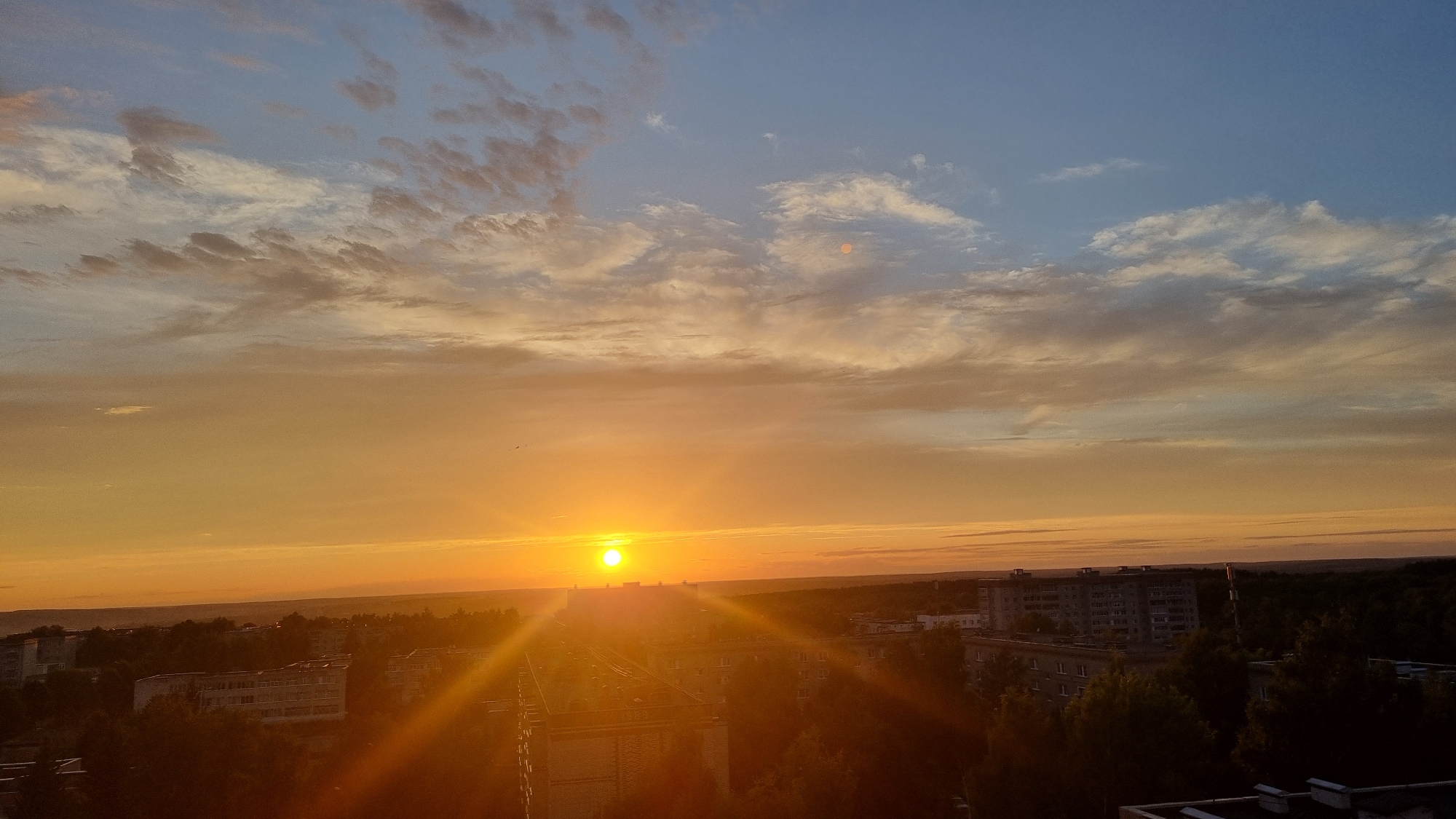 The image captures a sunset over the city of Lopatinka, Russia. The sun, a bright orange orb, is low on the horizon, casting long rays of light across the sky and the cityscape below. Wispy clouds, tinged with orange and pink, stretch across the sky, contrasting with the clear blue expanse above. The foreground is dominated by apartment buildings, some appearing weathered, interspersed with patches of trees and a sliver of railway track on the right. The overall impression is one of a typical urban evening, bathed in the warm glow of the setting sun.