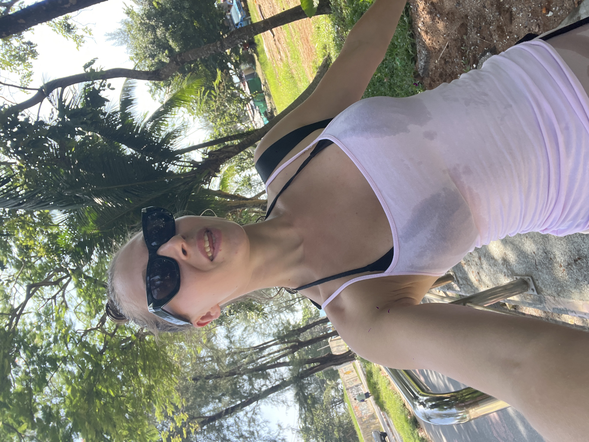 A young woman with her hair tied up in a bun, wearing black sunglasses, a light pink tank top, and a black bikini top, is taking a selfie outdoors. She has a backpack on her shoulders. The woman is standing on a dirt path with some rocks and greenery around her. In the background, there are palm trees, other trees with green foliage, and some buildings. To her left, there is a metal railing and a paved road with a car and two people walking in the distance. The scene appears to be in a tropical or subtropical location.