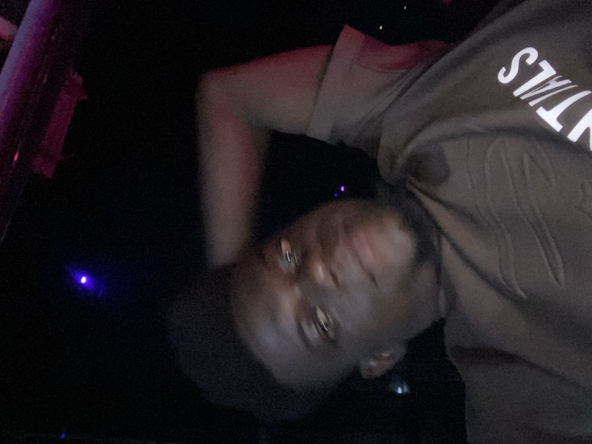 A dark-skinned person wearing a brown hooded sweatshirt with white letters is in the foreground. Their face is close to the camera. To the left is a dark, undefined area with a purple light source.