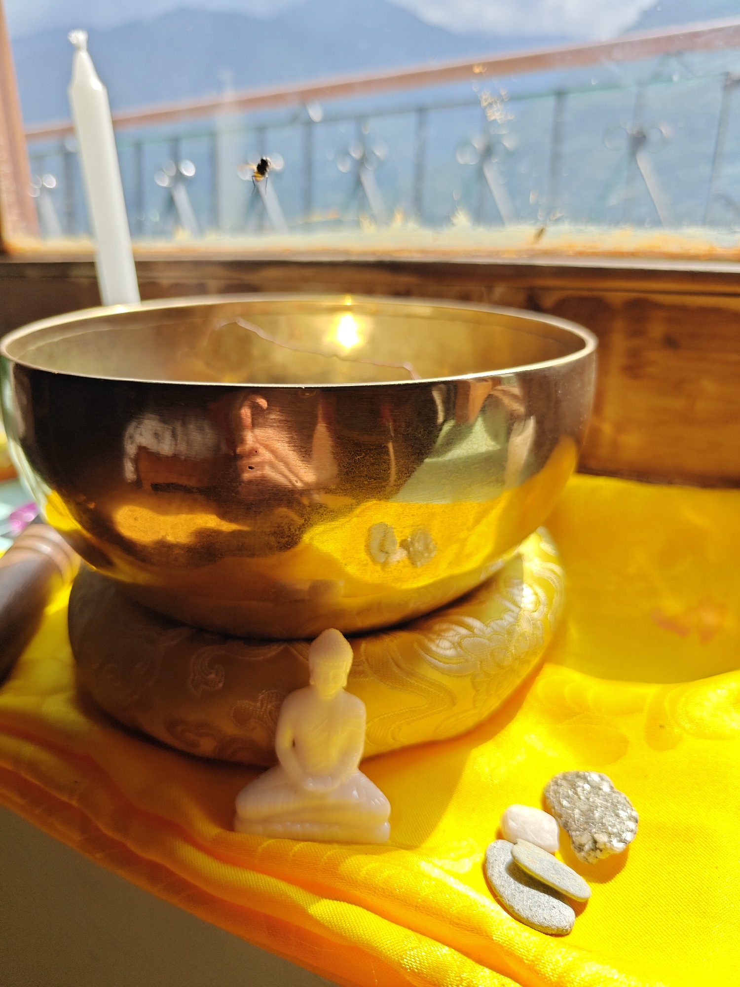 The image depicts a still life arrangement on a windowsill, likely in Vashisht, India. In the foreground, a golden singing bowl rests on a decorative cushion. To the right of the bowl, a small white figurine of the Buddha sits. A cluster of stones, including what appears to be pyrite, is also placed on the yellow cloth beneath the bowl. In the background, a white candle stands tall, and a small insect is visible on the windowpane, which looks out onto a hazy landscape with mountains and a body of water. The sunlight illuminates the scene, casting a warm glow and creating reflections on the singing bowl. The overall impression is one of tranquility and perhaps a spiritual or meditative setting.