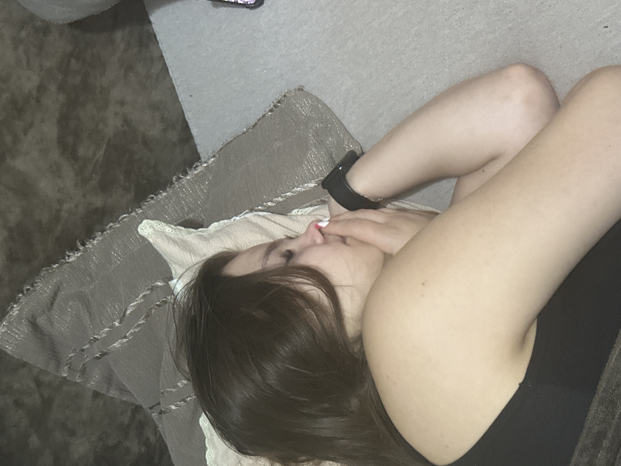 A young woman with dark hair is lying on her side, facing right, with her eyes closed. Her right arm is bent at the elbow, with her hand near her face. She is wearing a black tank top and a black smartwatch on her left wrist. The woman is resting on a light-colored pillow and a textured, grey blanket. The background appears to be a grey surface, possibly a couch or floor. There is no discernible information to determine the location within the city of Chișinău, Moldova.