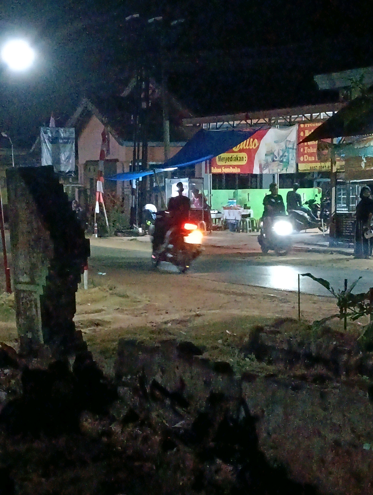In the city of Adirejo, Indonesia, at night, a street scene unfolds with a strong artificial light source illuminating the foreground from the left. In the center, two individuals ride motorcycles, their headlights cutting through the darkness. The motorcycle on the left is closer and its headlight is the brightest object in the image, casting a strong glow on the wet asphalt road. The rider is partially obscured by the light. To the right, another motorcycle carries a passenger, its headlight also visible. In the background, several buildings are dimly lit. One building features a banner with Indonesian text that reads "Menyediakan : Bahan Sembako" (Providing: Groceries). Another banner is partially visible. Small stalls or shops line the street, with some items displayed on tables. The ground appears to be unpaved in some areas, with patches of grass and debris. The overall impression is that of a bustling street market or commercial area at night.
