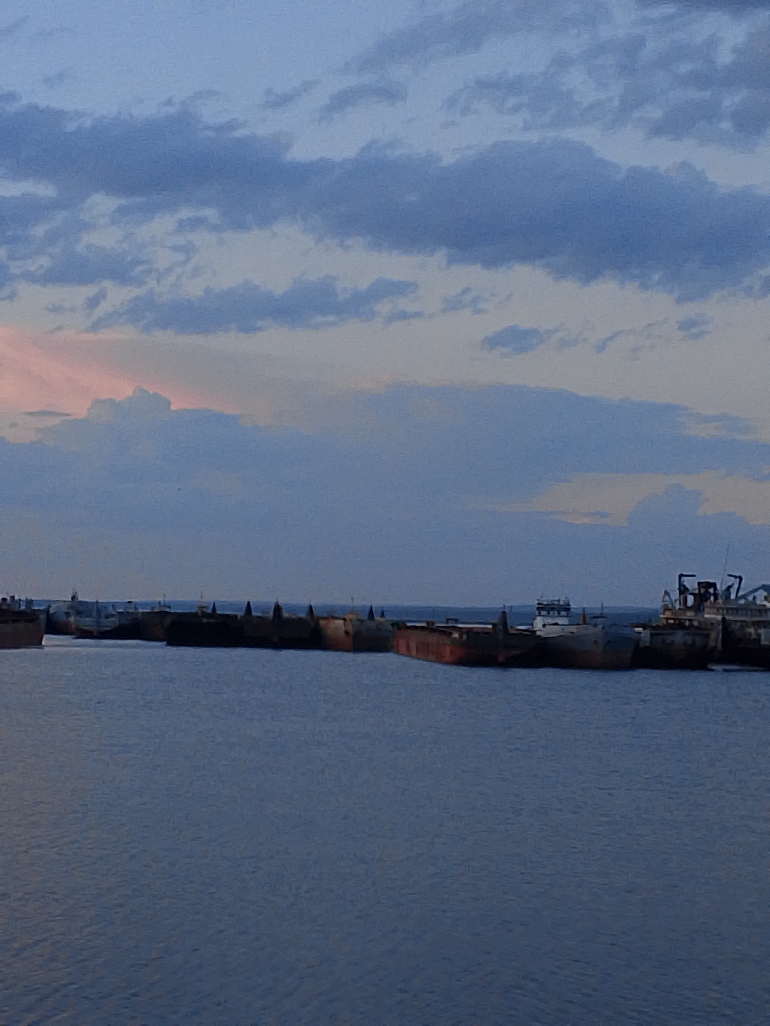 The image depicts a waterway with multiple boats lined up along the horizon, under a cloudy sky. The water is dark blue, and the sky is filled with blue and gray clouds, with some pinkish hues peeking through. The boats appear to be docked or anchored in a harbor. The scene has a muted, somewhat desaturated color palette.
While Chaikovsky, Russia is a potential location, the image alone doesn't provide enough information to conclusively verify it.