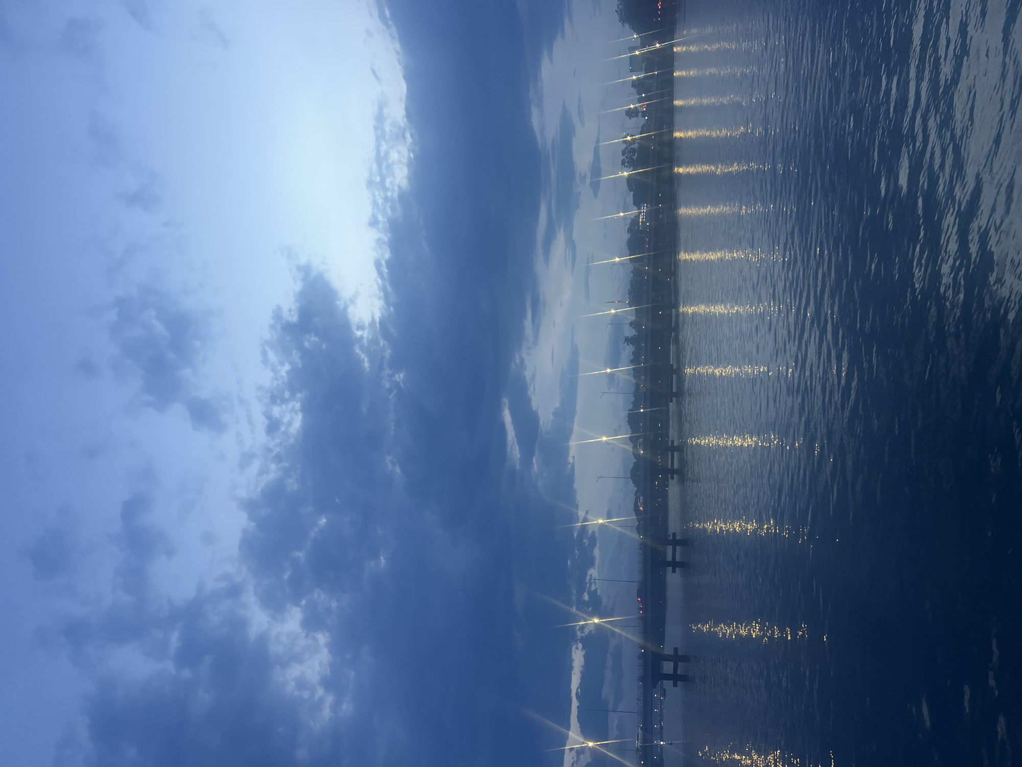 The image depicts a twilight scene over a body of water, likely a river or bay, in Thuận Hóa Ward, Vietnam. A bridge spans the water in the background, with traffic visible on its surface and streetlights illuminating its length. The water reflects the lights from the bridge and the sky, creating a shimmering effect. The sky is a deep blue, with scattered clouds, suggesting either early morning or late evening. The overall atmosphere is serene and tranquil.