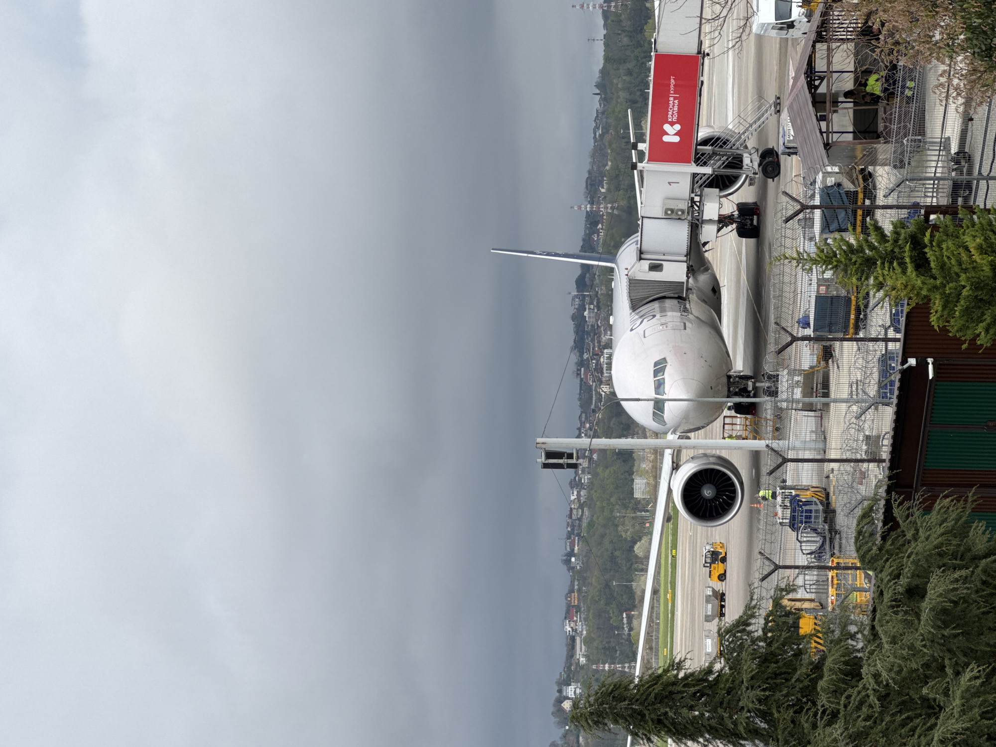 A passenger jet is docked at a gate at Sochi International Airport in Russia. The aircraft is white with dark blue accents on the tail. A jet bridge is connected to the plane. A red sign on the jet bridge displays the words "Красная Поляна" (Krasnaya Polyana) in Russian. Ground support equipment, including tugs and baggage carts, are positioned around the aircraft. Workers in yellow vests and safety gear are near the plane. The backdrop includes a cityscape with trees and buildings under a cloudy sky. The environment outside the airport has trees, buildings, and electric support towers, indicative of the nearby city, with overcast conditions.