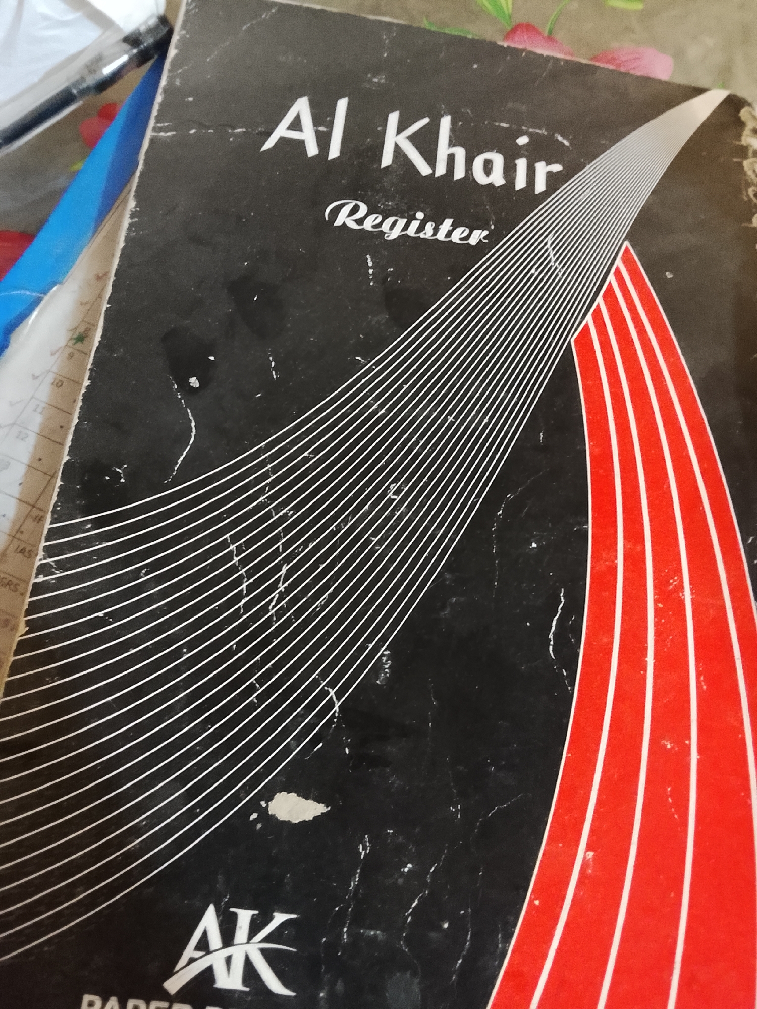 The image shows the cover of an "Al Khair Register." The register has a black background with the words "Al Khair" and "Register" printed in white. A design featuring white and red converging lines spans across the cover. The "AK Paper" logo is printed in the bottom left corner. The image does not offer sufficient information to confirm it was taken in Multan, Pakistan.