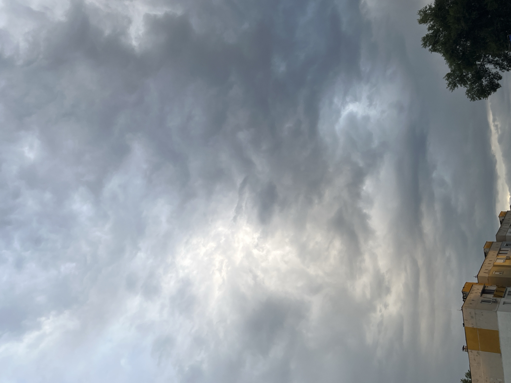 The image displays an expansive, heavily overcast sky, dominated by dense layers of gray and white clouds. The clouds exhibit varied textures, ranging from smooth, uniform gray areas to more turbulent, brighter patches where diffused light penetrates, hinting at a hidden sun. The overall impression is one of a very cloudy day, possibly preceding rain or depicting a generally somber atmosphere.
In the upper right corner, the dark green canopy of a tree is partially visible, its dense foliage contrasting with the lighter sky. Along the right side of the frame, a segment of a multi-story residential building can be seen. This structure features a blocky, apartment-style architecture, with exterior walls painted in shades of white and yellow. Signs of wear are apparent, including some discoloration and weathered surfaces. Visible details on the building include multiple windows and what appear to be air conditioning units installed on some balconies.
No people are present in the image, and no specific activities are taking place; it is a static landscape view. The time of day is daytime, though the heavy cloud cover obscures direct sunlight, suggesting either an early morning, late afternoon, or a perpetually gray day. The weather is distinctly cloudy. There is no visible text within the frame. This urban scene is set in Plovdiv, Bulgaria.