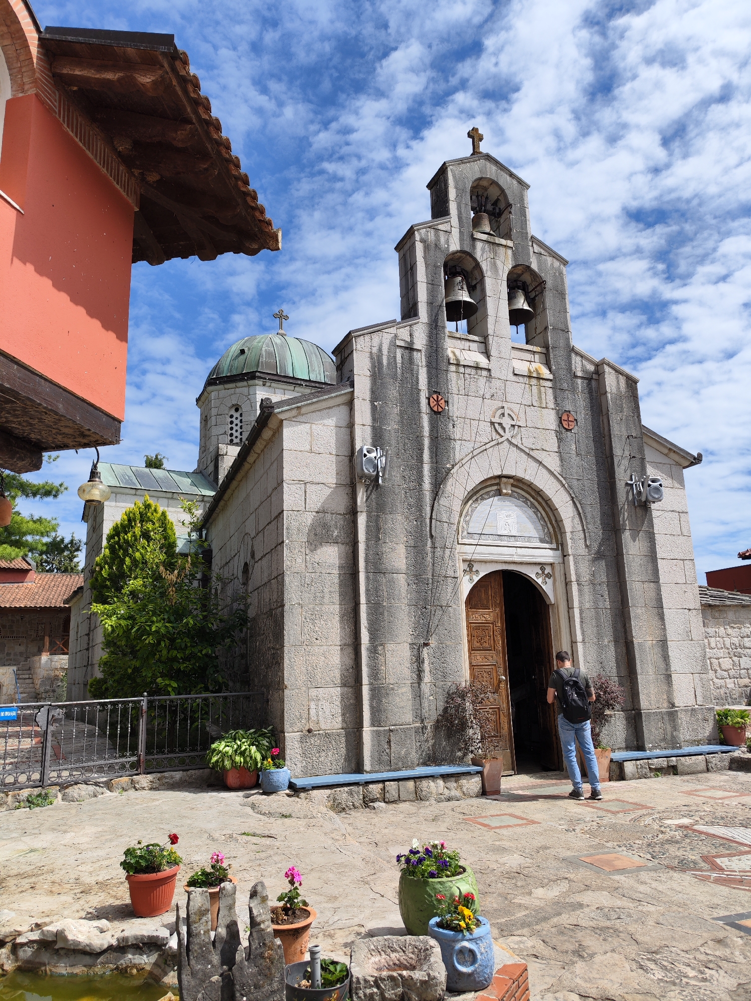 The image captures the exterior of the Hercegovačka Gračanica monastery in Trebinje, Bosnia and Herzegovina, on a bright, partially cloudy day. The monastery, built with light-colored stone, features a prominent bell tower topped with a cross, and a dome visible behind it. A man with a backpack stands at the open wooden doors of the monastery, seemingly about to enter. Several potted plants with flowers adorn the courtyard in the foreground, and a small stone-edged pond is also visible. A section of a building with an orange facade and dark wooden eaves is visible in the upper left corner. The courtyard has a paved surface with geometric patterns.