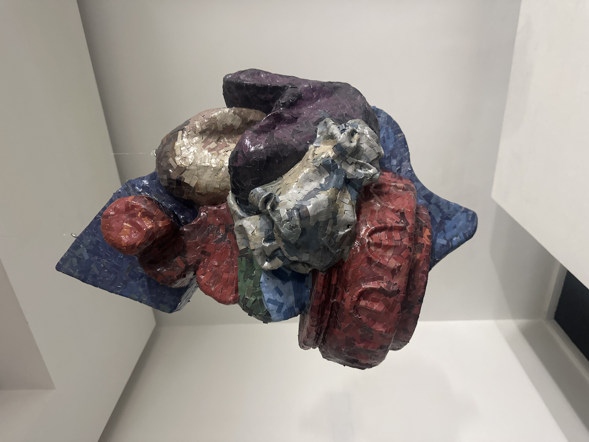 The image captures a vibrant, abstract sculpture suspended indoors, likely within an art gallery or museum in Buenos Aires, Argentina. The sculpture is characterized by its textured surface, composed of countless small, irregularly shaped pieces resembling a mosaic crafted from paper, fabric, or ceramic shards.
The artwork displays a rich palette of colors, including deep reds, various blues, purples, greys, off-whites, and touches of green. Its form is organic and complex, featuring several bulbous masses and draped elements. A prominent feature in the central right portion is a lighter grey and off-white section that strikingly resembles a human-like face, conveying an expression of distress or anguish with visible eye sockets and a downturned mouth. Other parts of the sculpture are more abstract and amorphous, contributing to the piece's overall dynamic and multifaceted appearance.
The sculpture is suspended by thin, nearly invisible wires against a plain, light-colored background of white walls and a ceiling, enhancing its visual impact. Subtle shadows are cast on the walls, suggesting directional lighting. No human activity or visible text is present in the scene. The overall impression is one of intricate detail, diverse textures, and a compelling interplay between recognizable forms and abstract shapes.