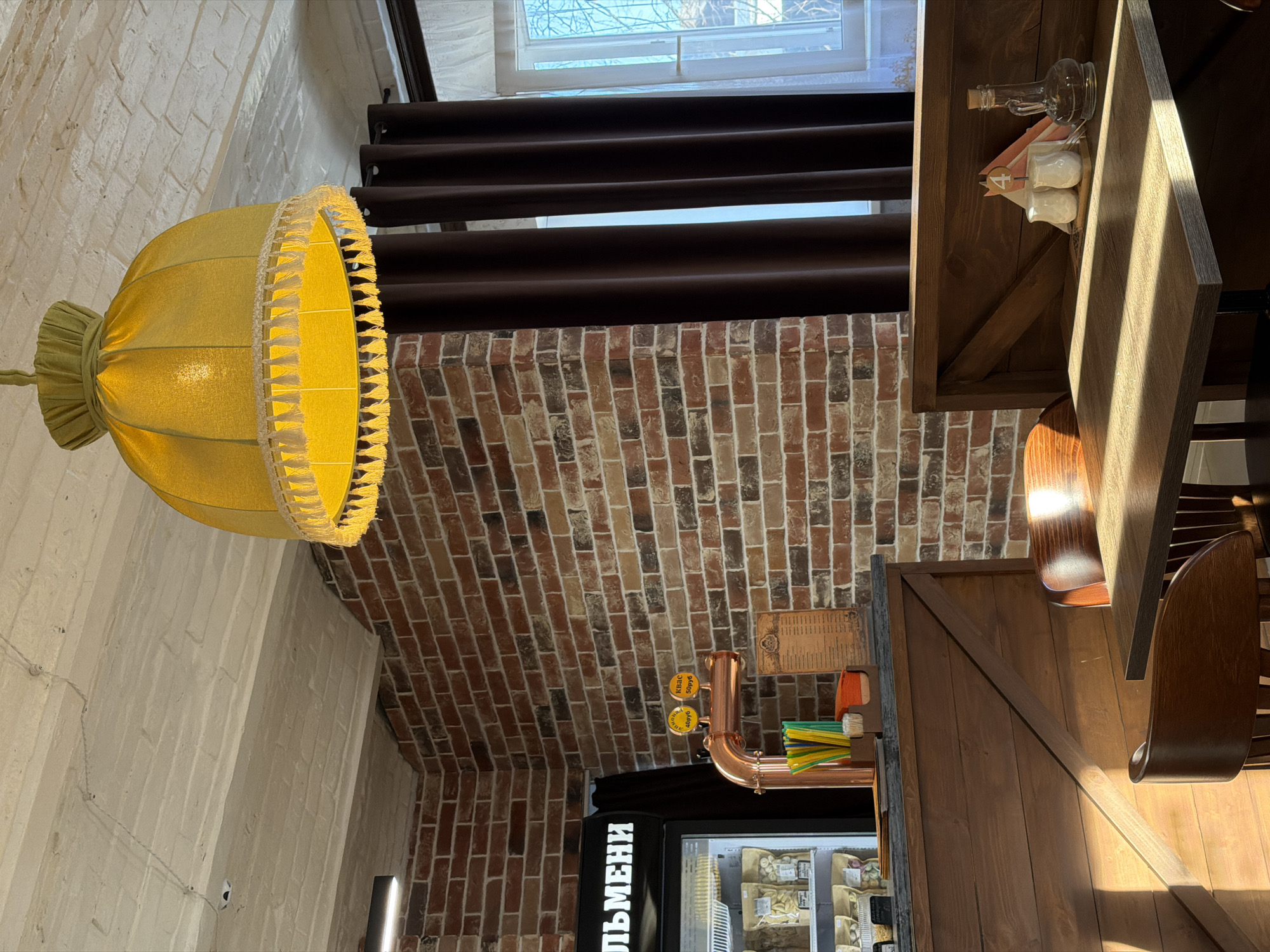 Here's a description of the image:
The image shows an interior scene, possibly of a restaurant or cafe, with a rustic aesthetic.
**Environment:** The space features a mix of brick and painted white brick walls, suggesting an older building. There's a window with dark curtains. Tables and chairs made of wood are present.
**Objects:**
* A large, yellow lampshade is a prominent feature.
* A beer tap is visible near a cooler that contains food.
* The label on the cooler is in Russian Cyrillic.
**Details:**
* The Cyrillic word "ЛЬМЕНИ" is visible on the cooler.
* There are condiments on the wooden shelf.
* A sign with Russian text and price is visible above the beer tap.
Given the Russian Cyrillic text, it's reasonable to infer that this photo was taken in Russia, though it is not possible to confirm it is in Sarapul with the available evidence.