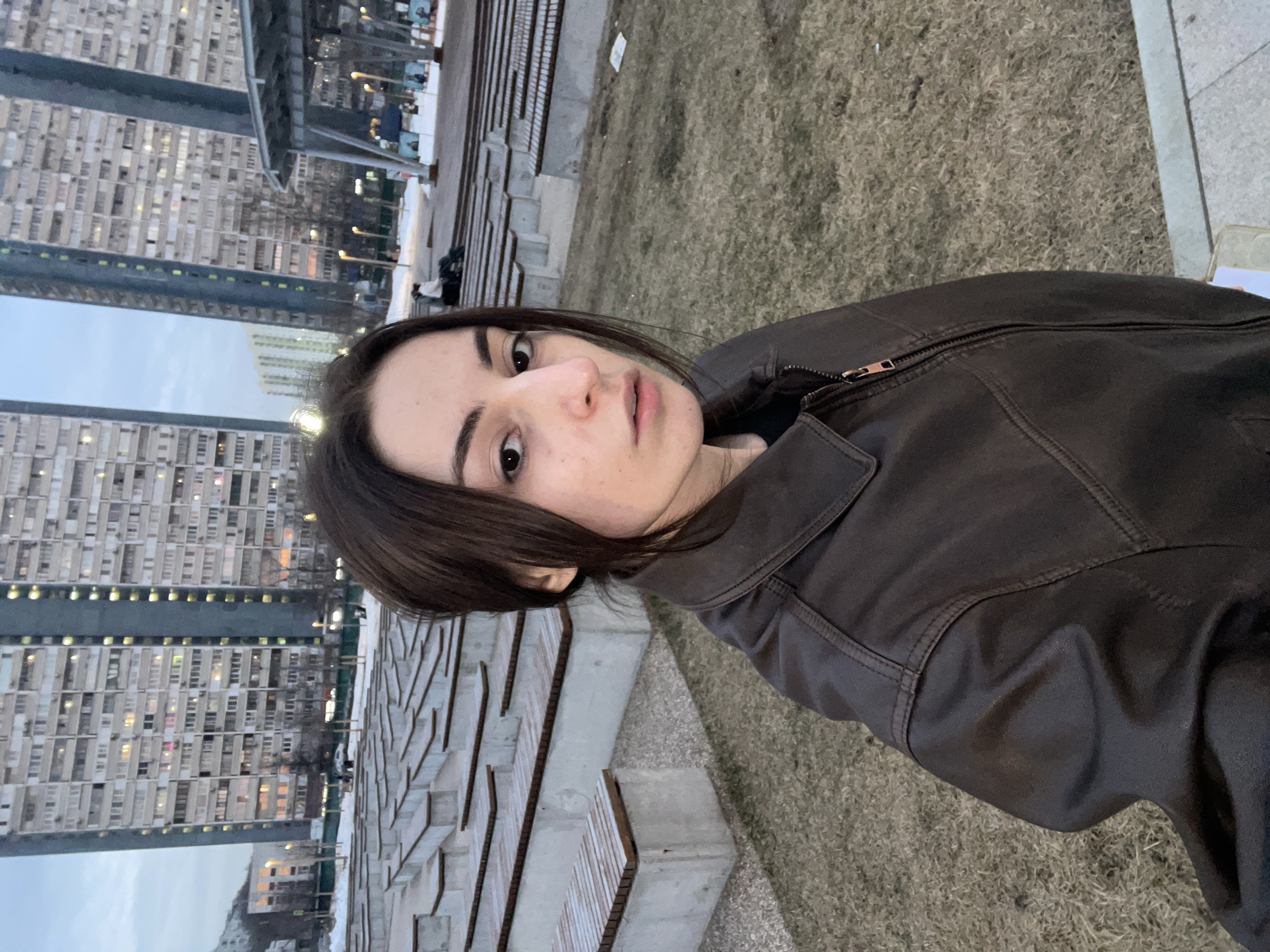 The image depicts a young adult female positioned in the foreground, facing the camera directly while capturing a selfie. She has dark hair pulled back and wears a dark brown collared jacket with a zipper, over a dark shirt. Her expression is neutral.
The scene is set in Khodynka Field Park (Khodynskoe Pole Park) in Moscow, Russia. The ground beneath her is covered with sparse, dry brown grass, indicating a late winter or early spring season. To the left and extending into the midground is a large, empty open-air amphitheater, constructed from concrete tiers topped with light-colored wooden planks.
In the midground, patches of snow are visible on the ground. To the right, part of a modern, grey multi-tiered architectural structure, possibly a pavilion, is present. Several bare trees are scattered throughout the park. Distant figures are visible, including one seated on a bench in the midground, and others further away near the modern structure. A faint outline of a light-colored vehicle is discernible in the far background on a road.
Dominating the far background are two tall, multi-story residential buildings characteristic of Soviet-era panel construction (e.g., P-44 series), with numerous windows, many of which are illuminated with warm light. The sky above is a light blue-grey, suggesting late afternoon or early evening.