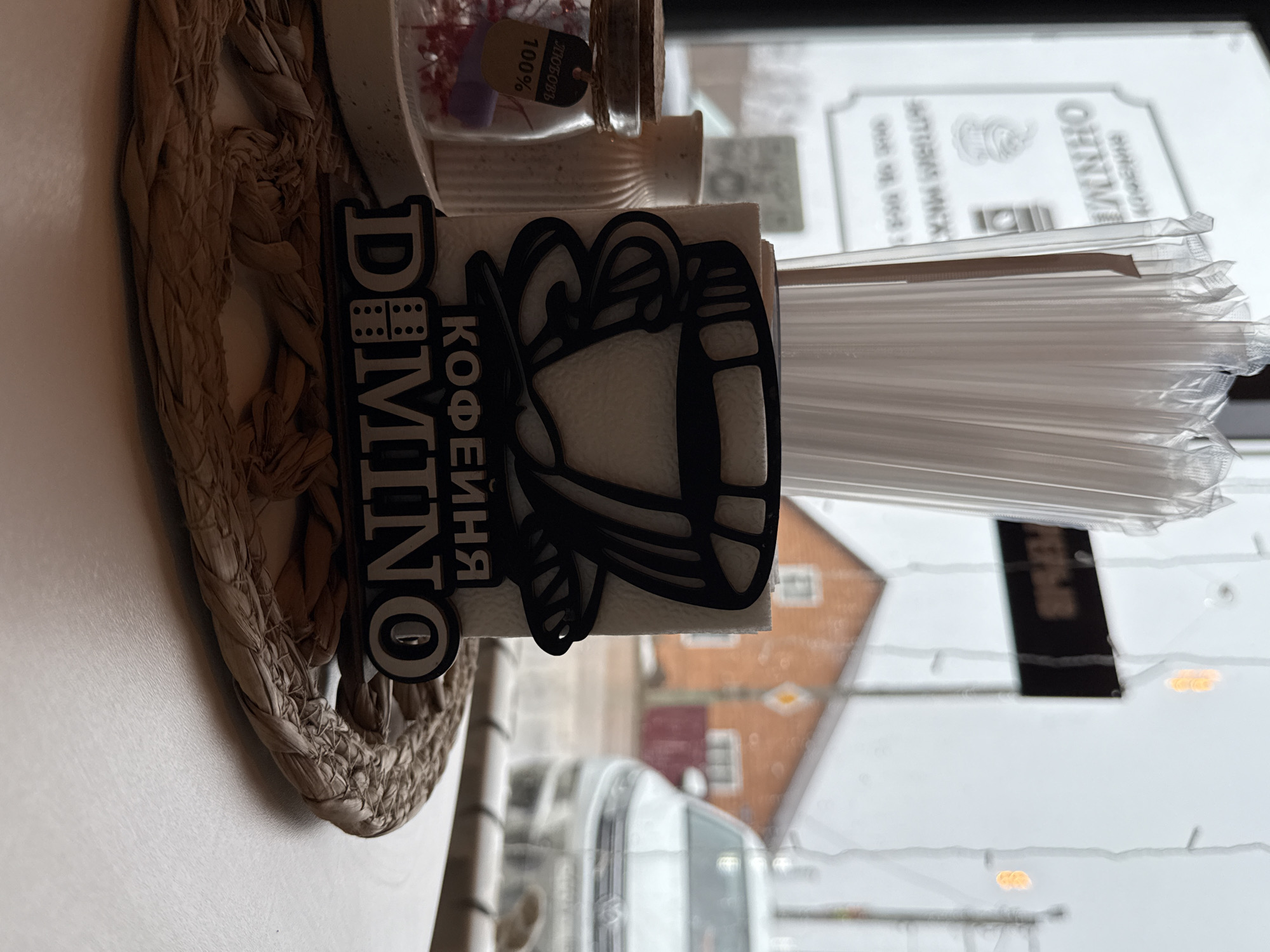 Here is a description of the image.
The image shows a table setting near a window, with elements indicating the scene is in or associated with the city of Osa, Russia. The table setting includes a woven placemat, a napkin holder with a coffee-themed design and the inscription "DOMINO" and "КОФЕЙНЯ" (coffee shop in Russian), a container of small items, and a dispenser with straws. Through the window, a blurred view of buildings and possibly streets is visible, suggesting an urban environment. A sign with the name "OHHNO" is noticeable in the background. The general impression is a snapshot of a cafe or restaurant setting in Osa.