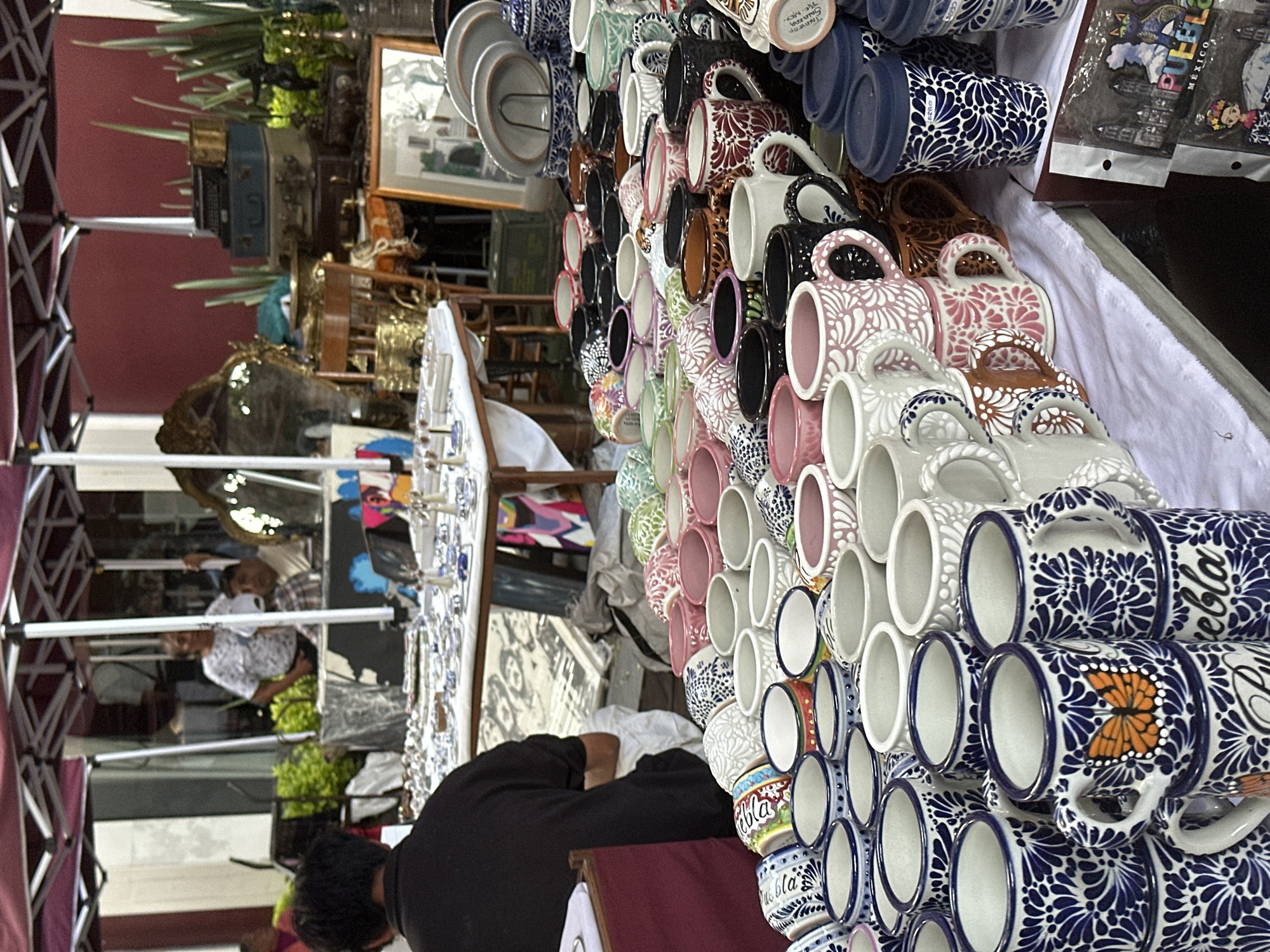 The image captures a bustling marketplace scene, potentially in Puebla, Mexico, judging by the presence of "Puebla" branded mugs. An array of handcrafted pottery mugs in various colors and designs, including Talavera style, are prominently displayed on a white-draped table. A person dressed in black stands behind the display, possibly tending to the merchandise. Another table is positioned to the left, featuring an assortment of jewelry. In the background, there are more goods for sale. Other people are visible, browsing or walking through the marketplace. The setting appears to be an outdoor market, likely a tourist spot.