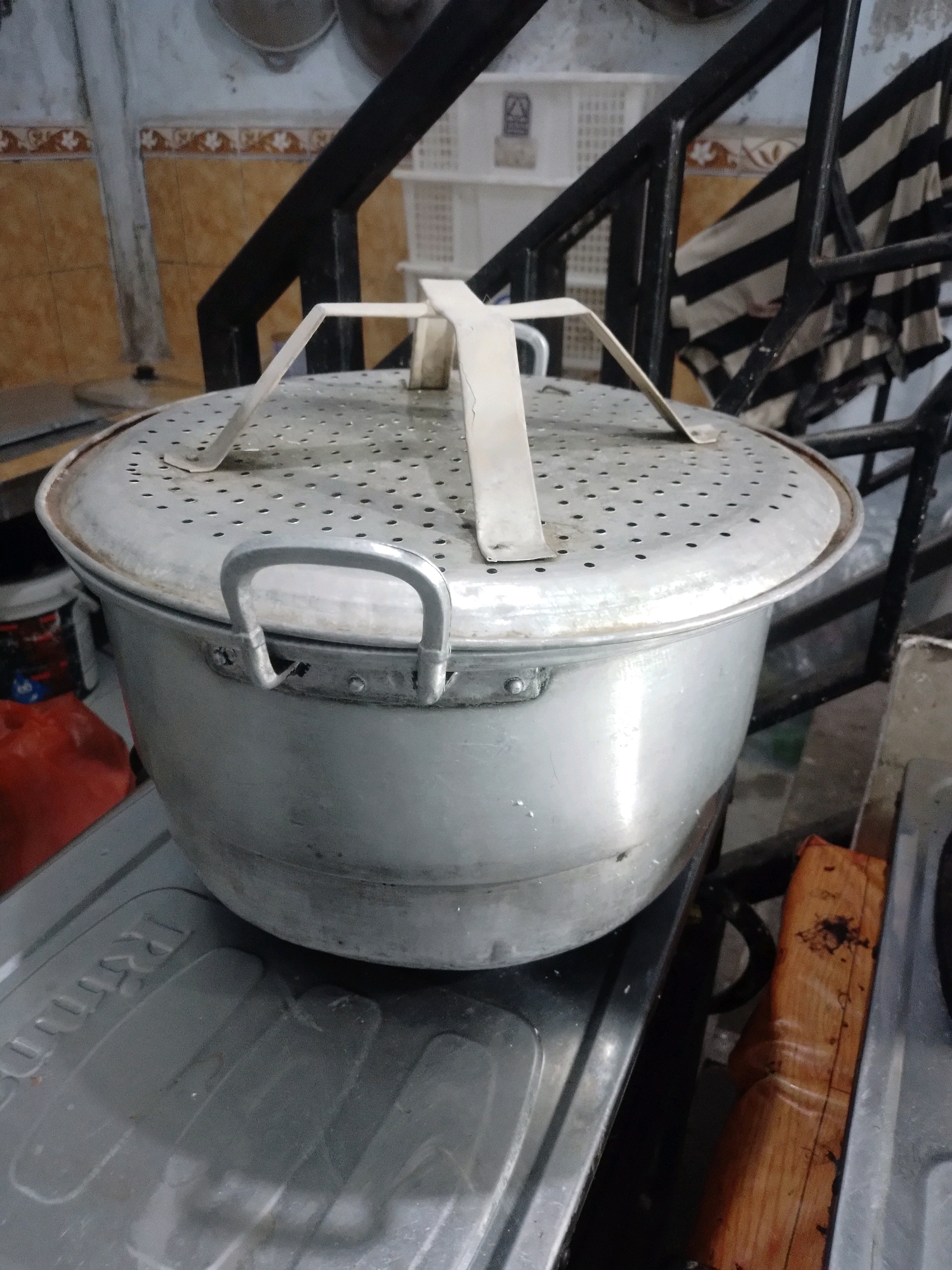 A large, aluminum pot with a perforated lid and two side handles sits on a gas stove in a kitchen. The pot is the primary subject, appearing well-used. The lid has a central, U-shaped handle and numerous small holes. The pot itself is round and appears to be made of a polished aluminum alloy.
The setting is a kitchen, evident from the presence of the gas stove, the tiled backsplash, and what appears to be a metal railing behind the pot. In the background, a rack with black and white striped fabric is visible, possibly a dishtowel or apron. Further back, a white plastic crate and some ceramic tiles with a floral pattern can be seen.
There is no activity taking place in the image; it is a still shot of the pot on the stove. The lighting suggests it is daytime, likely indoors with artificial or natural light.
On the surface of the stove in front of the pot, there is embossed text that is partially visible and difficult to read. There is also a piece of wood with dark stains on it, placed on the stove next to the pot. The overall impression is of a functional, lived-in kitchen.