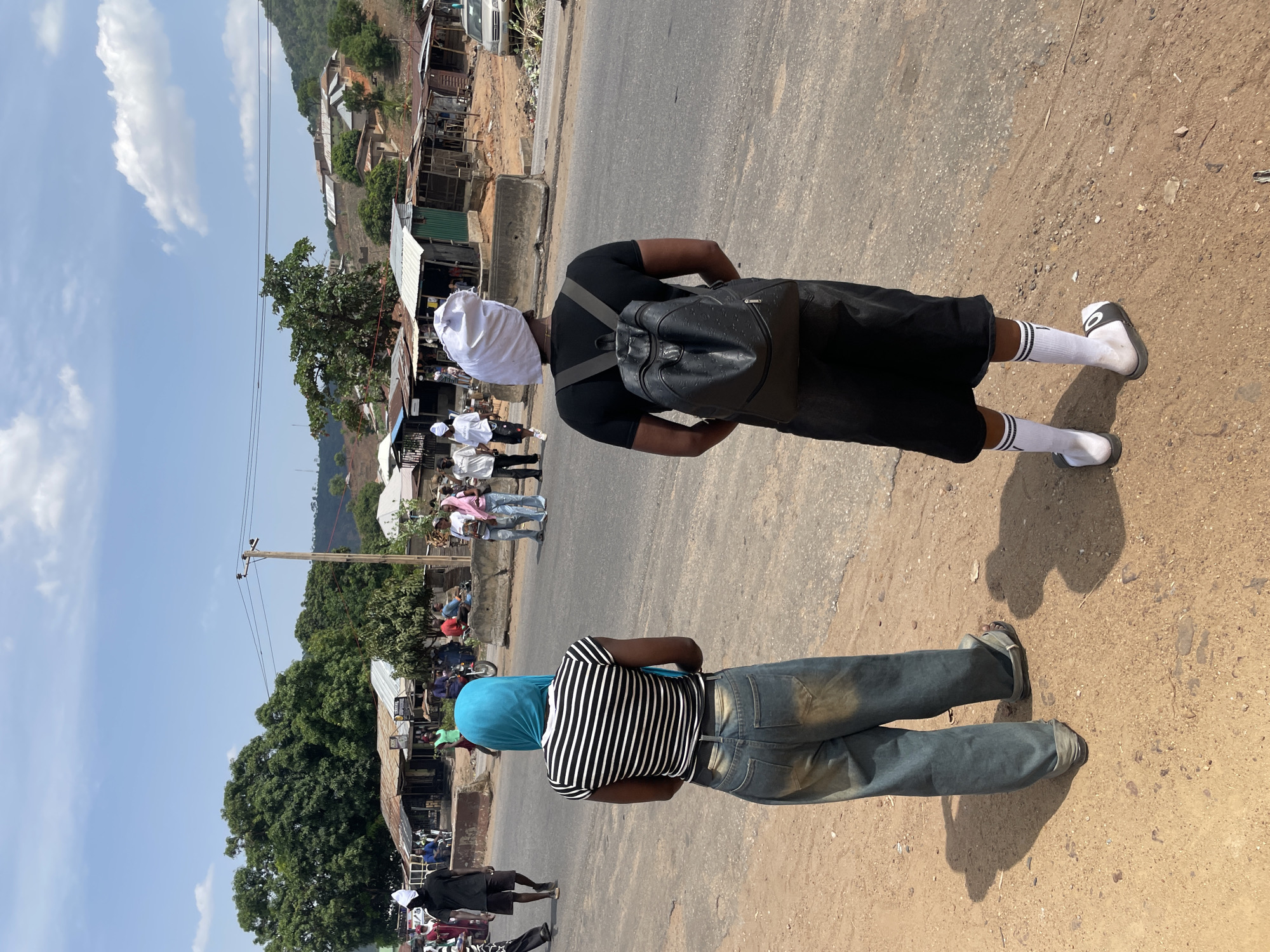 Two people are standing on a road, one wearing a white head covering, a black shirt, black shorts, white socks with black stripes, and black sandals, and carrying a black backpack, and the other wearing a blue head covering, a striped shirt, and blue jeans. They are facing away from the camera and appear to be on the outskirts of the city of Lokoja, Nigeria, based on the presence of informal structures and other people milling about nearby.