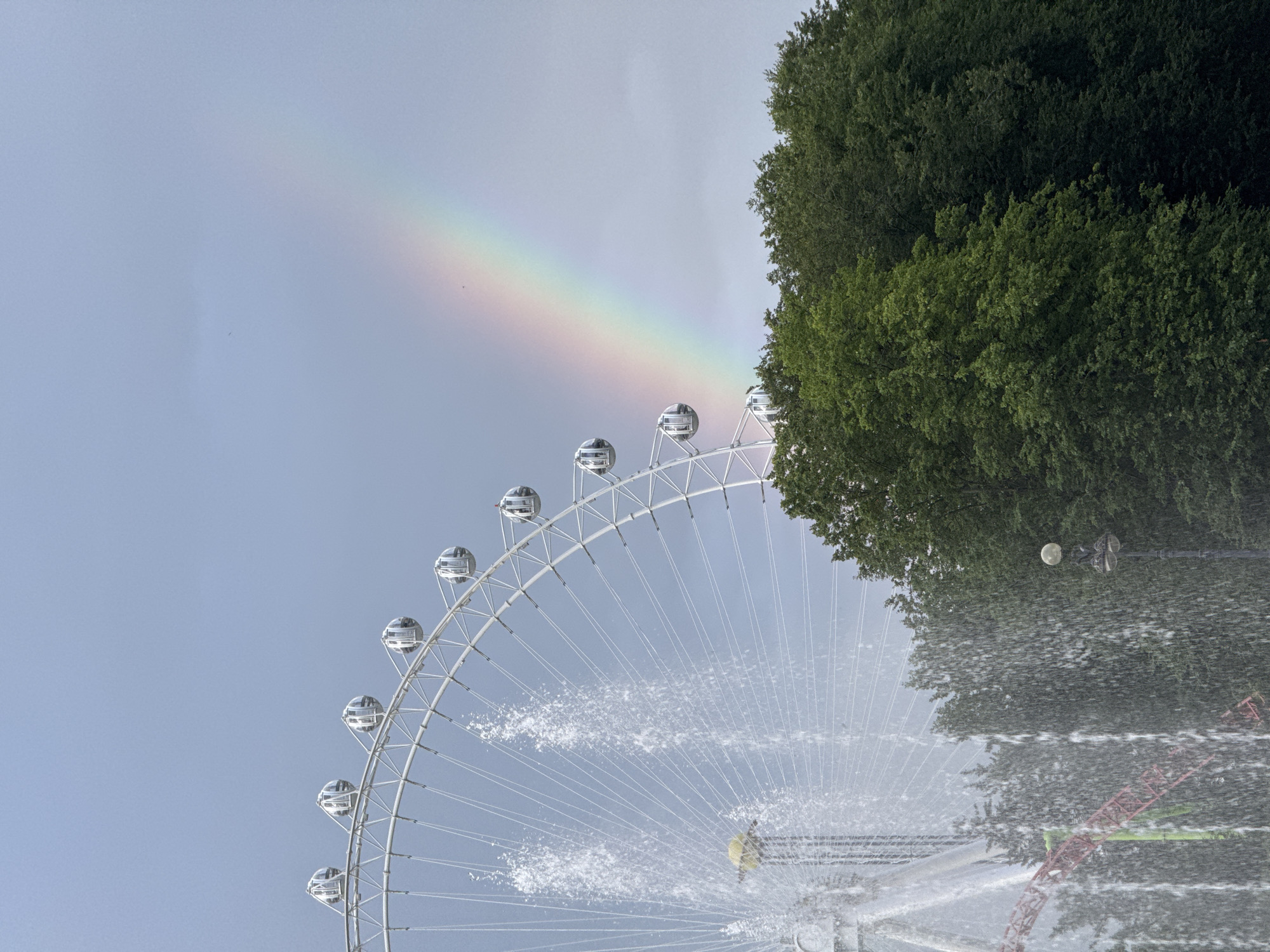 The image depicts a ferris wheel and a fountain in Saint Petersburg, Russia. The ferris wheel is white and features multiple enclosed cabins. A prominent fountain is spraying water in the foreground, partially obscuring a section of the ferris wheel and a roller coaster track. Lush green trees form a dense backdrop to the scene. A rainbow arcs across the overcast sky to the right of the ferris wheel. The lighting suggests it is daytime, likely after or during a rain shower.