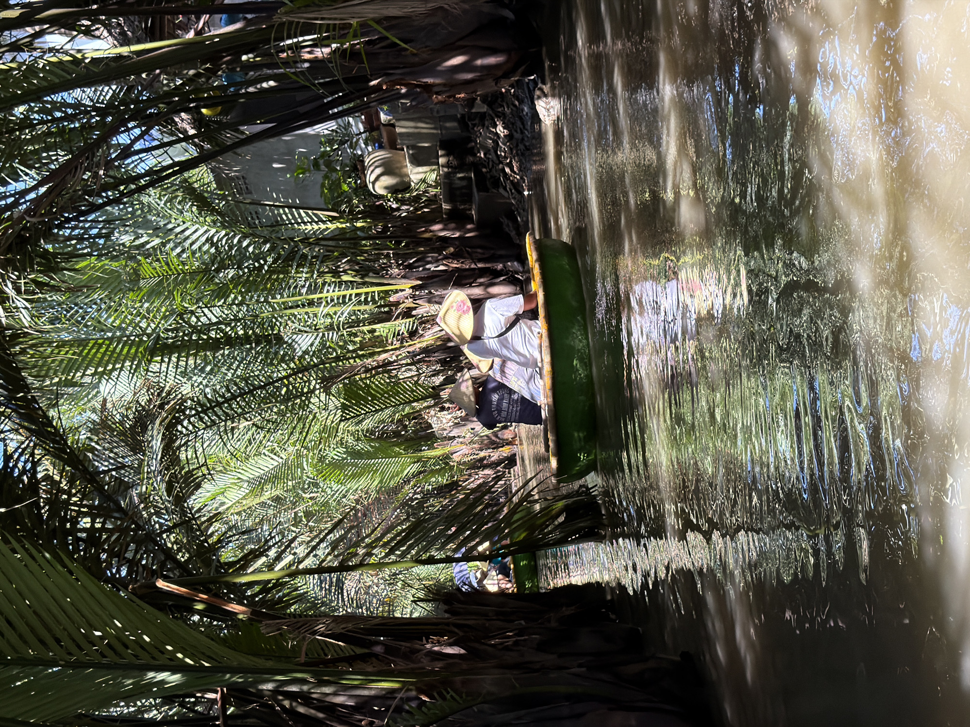 A boat tour is underway along a narrow canal surrounded by dense, lush palm trees. The water is murky and reflects the green foliage and sunlight. Two people are visible in a small, round, green boat in the foreground. Both are wearing traditional conical hats. The person in the back is wearing a black shirt with white text and the person in front is wearing a white shirt. Another boat with two people is partially visible further down the canal on the left. In the background, there are structures and more dense vegetation, suggesting a rural or semi-rural area within Đà Nẵng, Vietnam, likely a tourist attraction showcasing the natural waterways and local life.