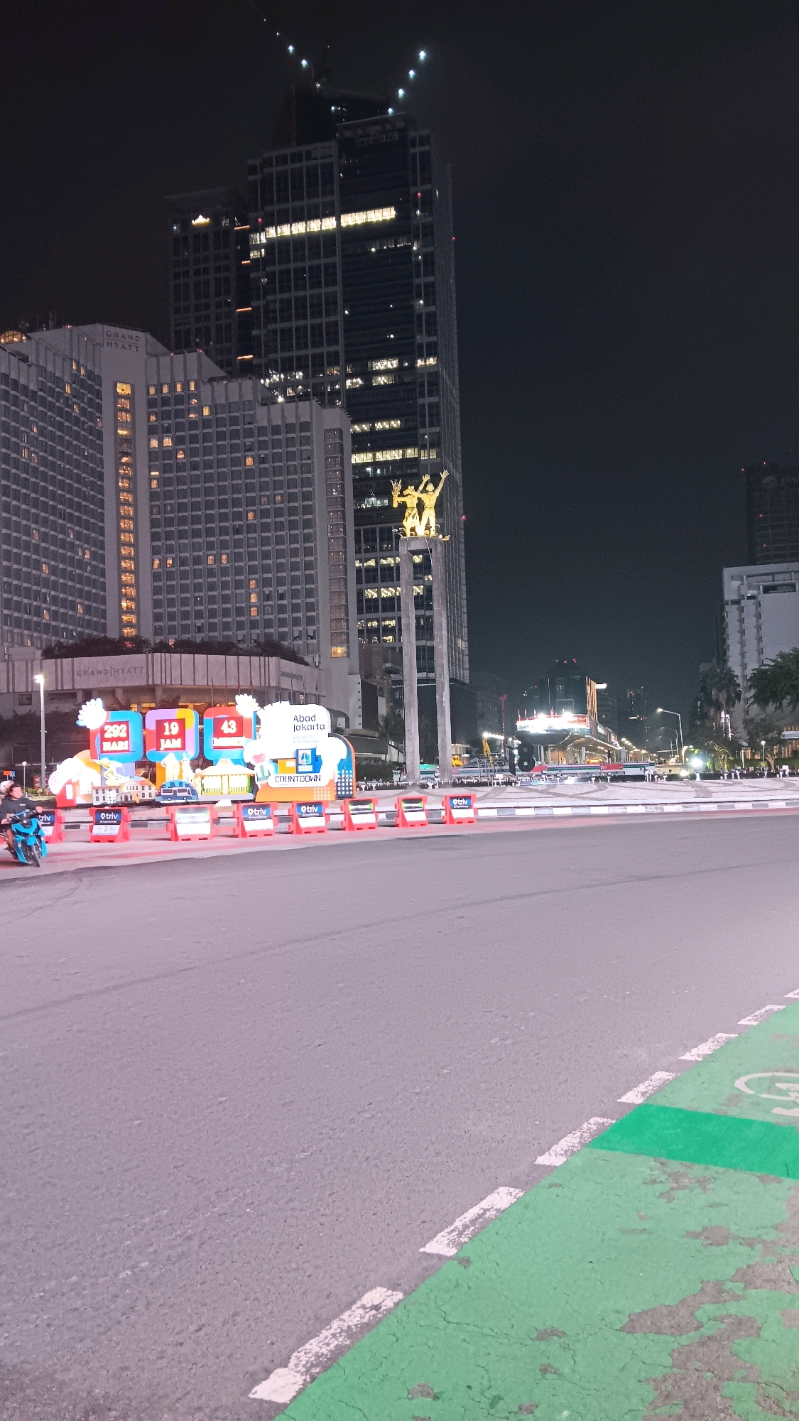 This is a nighttime street-level photograph of the Monas area in Central Jakarta, Indonesia. The image is dominated by the facade of the Grand Hyatt hotel on the left and a tall, modern skyscraper on the right, possibly The Plaza office tower, which is under construction or renovation at the top. In the mid-ground, there's a large, illuminated digital countdown display with the text "Abad Jakarta" and "COUNTDOWN." The display features numbers like "292," "19," and "43" along with graphics of buildings and vehicles. A motorcycle with two occupants is visible on the left side of the frame, partially obscuring a row of orange traffic cones and barriers. The street in the foreground is paved, with a green-painted lane marked by white dashed lines. The overall ambiance is urban and dynamic, characteristic of a busy city at night.