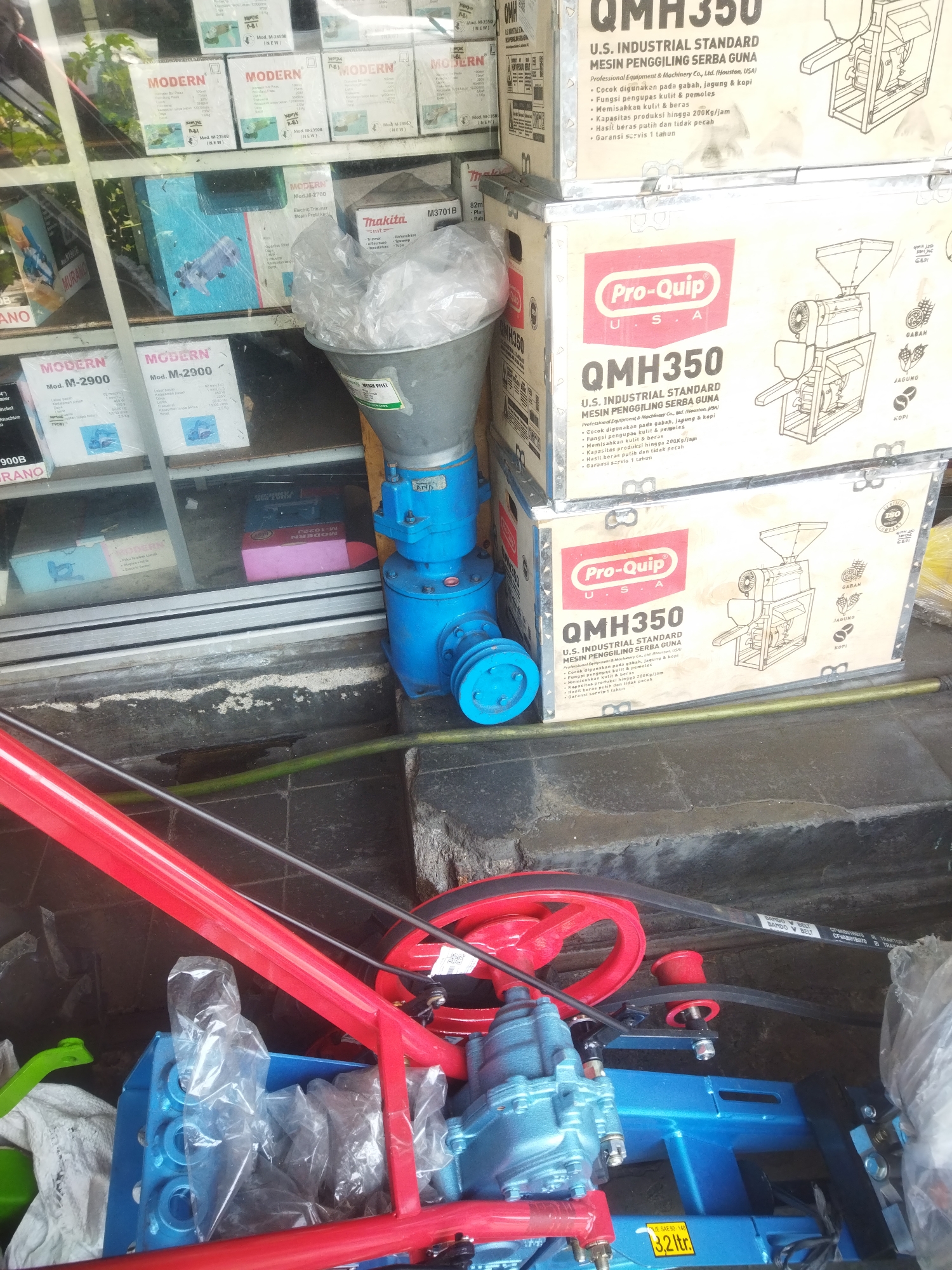 In Giripurwo, Indonesia, a close-up shot showcases agricultural machinery and its packaging, likely outside a retail establishment. The scene is dominated by a bright blue and red milling machine, partially obscured by plastic packaging. The machine features a prominent red wheel and a black drive belt system. Surrounding the machine are cardboard boxes, some displaying branding such as "MODERN" and "Pro-Quip QMH350 U.S. INDUSTRIAL STANDARD MESIN PENGGILING SERBA GUNA." These boxes indicate the machinery is designed for grinding various grains like gabah (paddy), jagung (corn), and kopi (coffee). The background reveals a store interior with shelves stocked with more product boxes, some featuring images of the machinery within. A green hose lies on the ground near the machine. The overall impression is one of commercial activity, with new machinery awaiting sale or use.