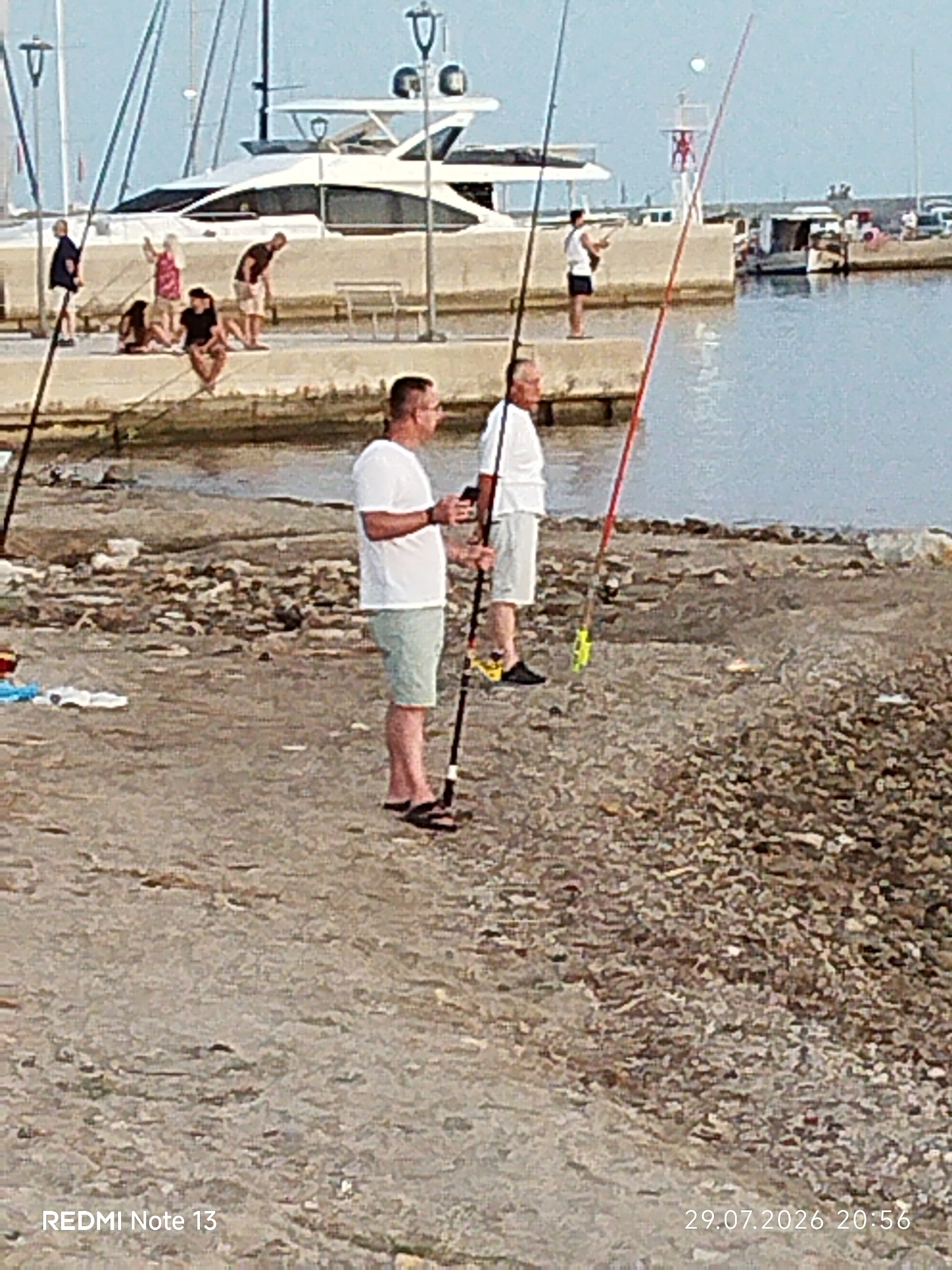 The image shows a waterfront scene in Sithonia Municipal Unit, Greece. Two men are fishing in the foreground. The man on the left is wearing a white t-shirt and light-colored shorts, holding a fishing rod. The man on the right is wearing a white t-shirt and darker shorts, also holding a fishing rod. In the background, several people are visible on a pier near a large white yacht. The environment appears to be a harbor with calm water. The ground in the foreground is covered with pebbles and sand. A red fishing rod is prominently displayed, leaning against a textured surface. The bottom left corner displays "REDMI Note 13" and the bottom right corner displays "29.07.2026 20:56".