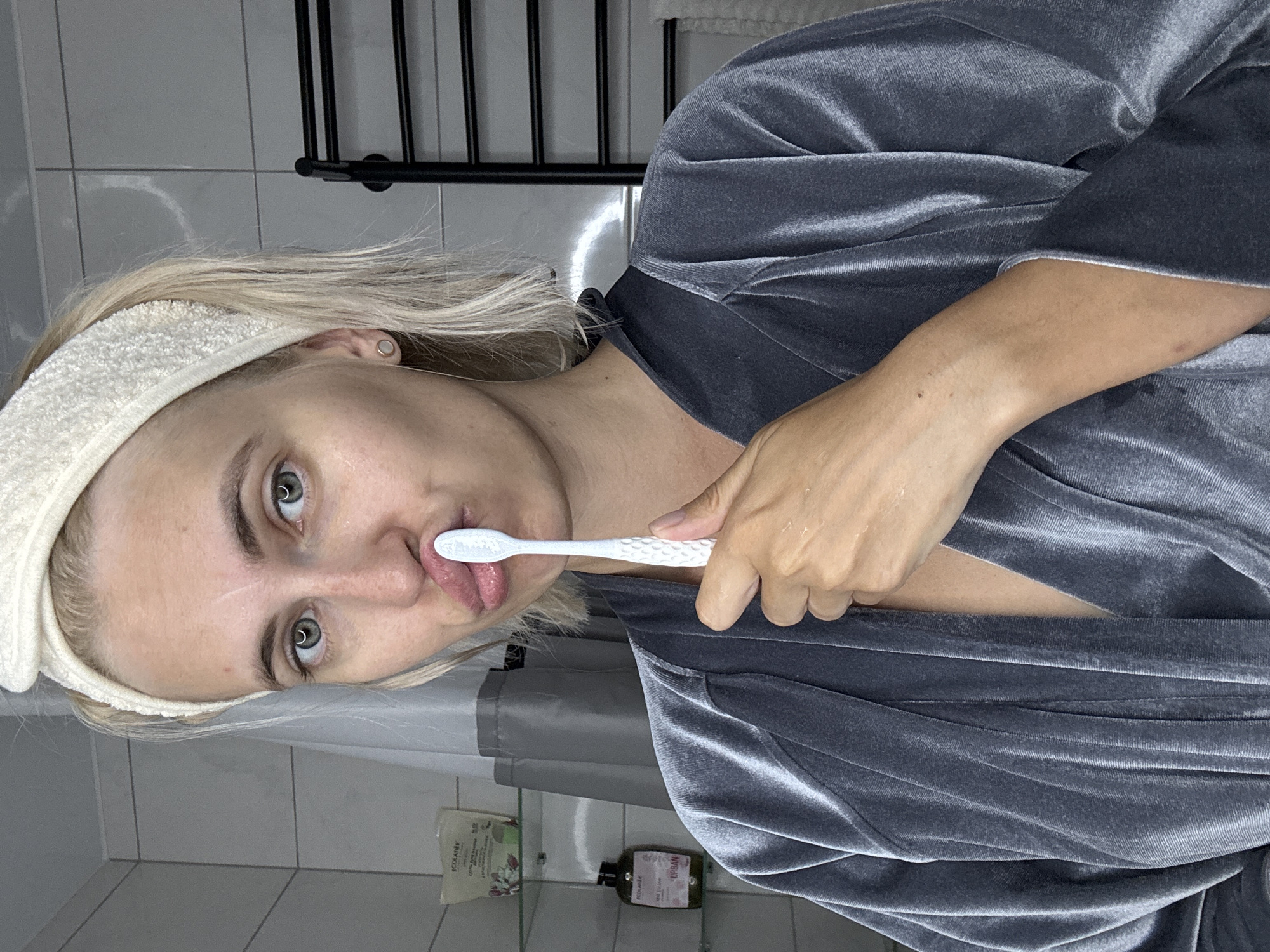 A blonde woman with a white headband and a grey velvet robe is holding a white toothbrush to her lips, making a pouty face. She is indoors, likely in a bathroom, with tiled walls and a shower curtain visible in the background. A black towel rack is also present. The woman appears to be preparing to brush her teeth.