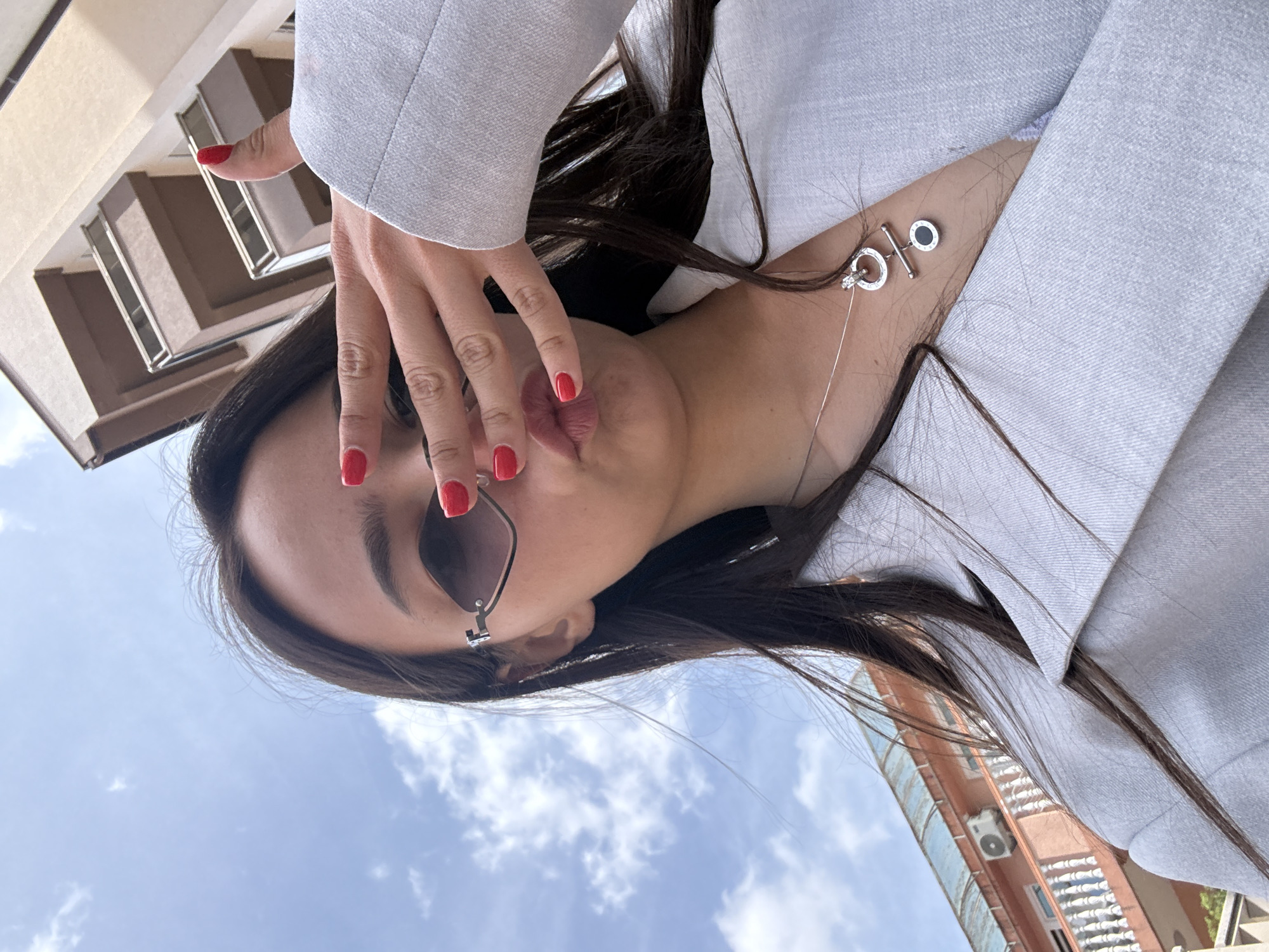 A woman is seen from a low angle, potentially holding the camera herself, against a backdrop of a city environment. She is wearing a light-colored blazer, and the open neckline reveals a delicate silver necklace with distinctive circular and t-bar pendants. One hand is raised near her face, fingers gently touching her lips, and her nails are painted a vibrant red. She is wearing sunglasses with a minimalist frame.
In the background, portions of buildings are visible against a blue sky dotted with scattered clouds. One building appears to have apartments with balconies and air conditioning units, while another shows architectural details with windows.