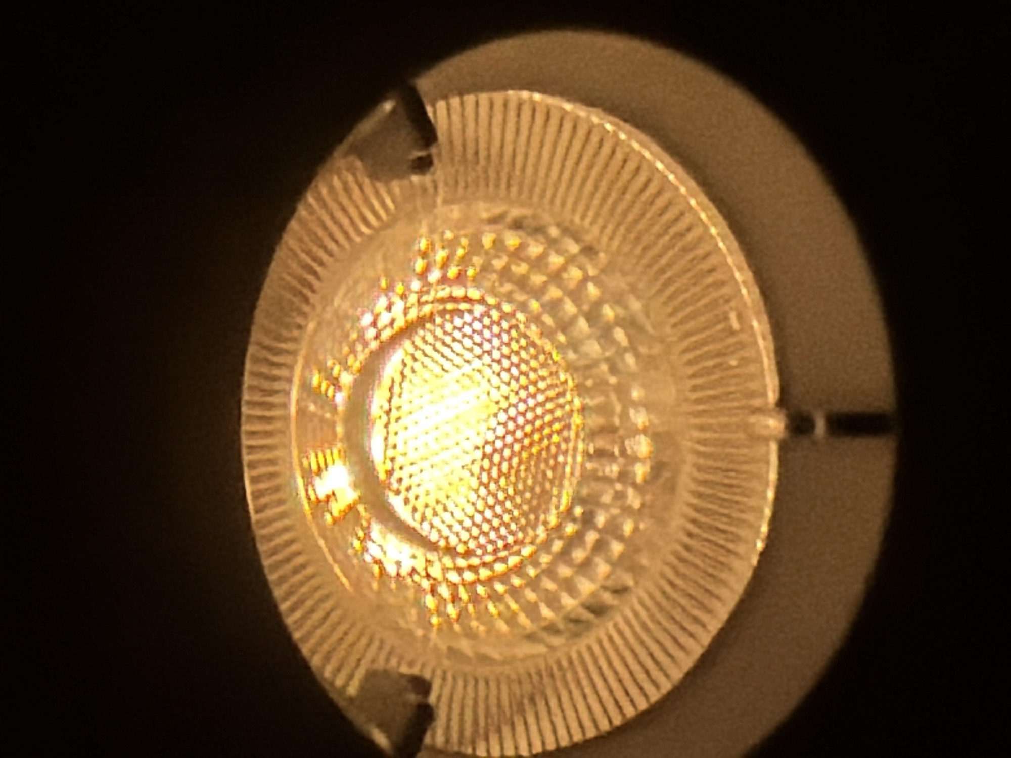 This image, taken in Maidstone, United Kingdom, presents a close-up, top-down view of an illuminated artificial light source. The central element is a circular array of numerous small, individual light-emitting diodes (LEDs) configured in a honeycomb-like pattern, emitting a warm, golden-yellow light. Surrounding this central luminous area is a clear, circular diffuser or lens featuring concentric rings and distinct radial fluting or grooved patterns designed to refract the light. The outer rim of the fixture is composed of a dark, matte, brownish-gray material, likely the housing. Three dark, narrow securing clips or mounting points are visible, positioned at approximately the 10 o'clock, 2 o'clock, and 6 o'clock positions relative to the light fixture. The immediate environment surrounding the light fixture is uniformly black, indicating a dark setting or focused illumination. No people, discernible actions, or interactions are visible within the frame.