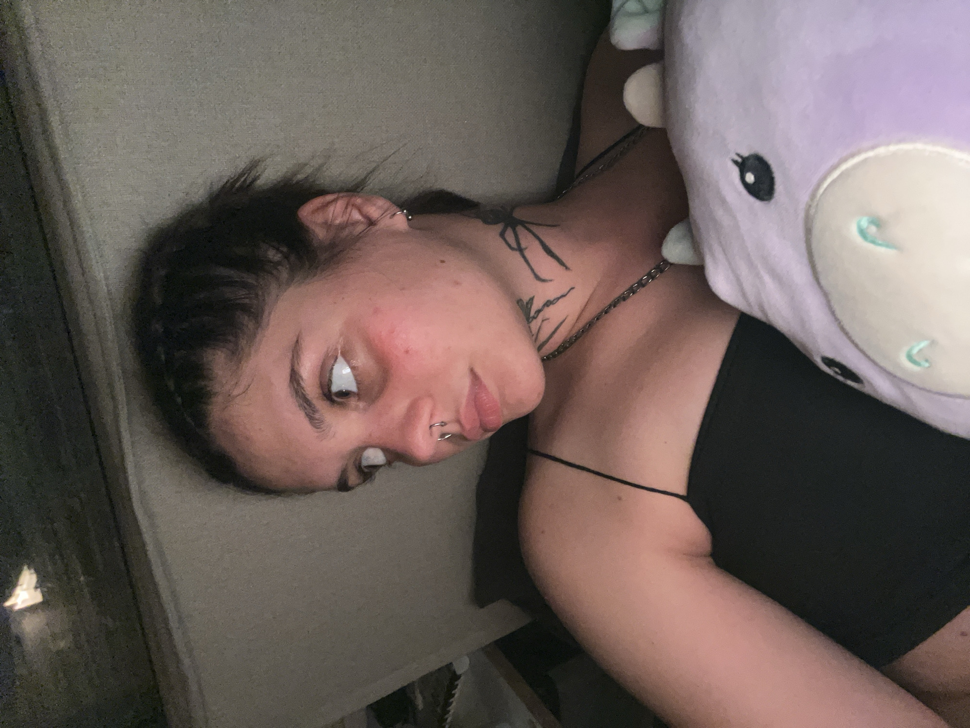 The image shows a close-up, low-angle portrait of a young woman with dark hair pulled back, wearing a black spaghetti strap top. She has multiple piercings, including a septum ring and an earring. There are tattoos on her neck, one appearing to be a spider. She is holding a purple stuffed animal, possibly a cow or dragon, with visible eyes and ears. The woman is looking to her left, with rosy cheeks and her hair slightly disheveled. The background appears to be indoors, with a textured surface and some lighting that suggests a dimly lit environment. There is no visible indication of the city of Genemuiden, Netherlands, in this image.