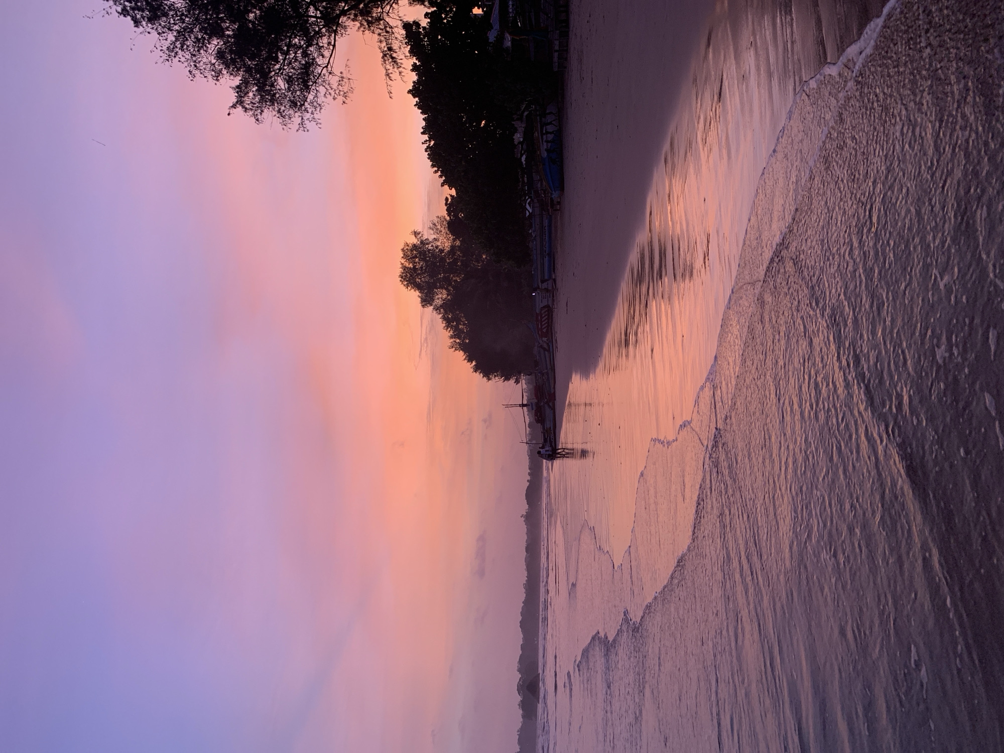 The image depicts a serene beach scene in Weligama, Sri Lanka, at dusk. The sky is a gradient of soft purples and pinks, transitioning to a warm orange near the horizon. Gentle waves roll onto a sandy shore, their shallow waters reflecting the colors of the sky.
In the mid-ground, two figures are walking along the wet sand. Further down the beach, several small fishing boats are pulled ashore, silhouetted against the vibrant sky. Lush green trees and foliage line the edge of the beach, creating a natural border. The overall atmosphere is peaceful and tranquil, characteristic of a tropical sunset.