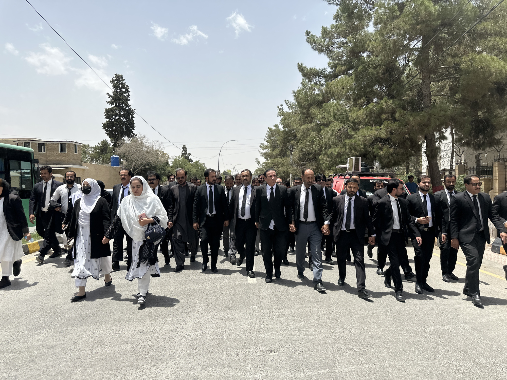 In the city of Quetta, Tehsil, Pakistan, a group of people, predominantly men in dark suits and ties, are walking on a paved road under a bright, cloudy sky. The group appears to be a mix of legal professionals, suggested by their attire. A few women are also present, one in a black and white ensemble and another in a white hijab and a white outfit, holding a pink cell phone and a black handbag.
The environment shows a street with some trees and buildings in the background. A large green bus is partially visible on the left side of the frame. A red vehicle, possibly a fire truck, is also present behind some of the individuals. The road has faint white lines, indicating it is a marked street. The overall scene suggests a procession or a march, with the individuals moving forward together.
