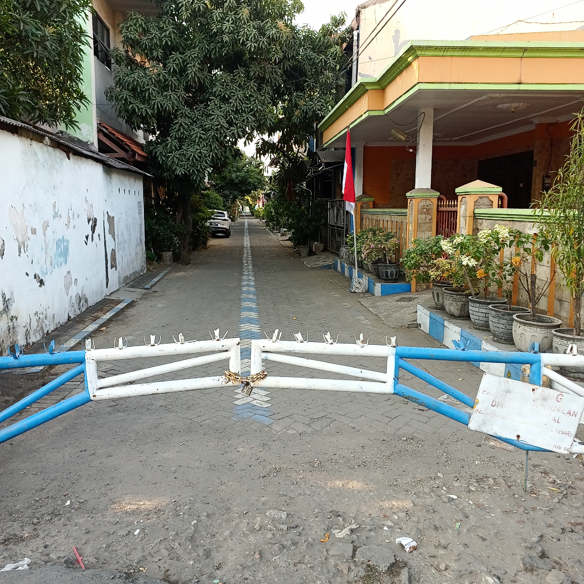 In Wedoro, Indonesia, a gated street is visible from the perspective of the barrier. The barrier, constructed of white and blue metal bars, is secured with a chain and two padlocks, blocking access to the street. The street is paved with interlocking bricks and marked with a dashed blue line down its center. A white car is parked on the left side of the street in the distance. On the right side of the street, a house with an orange facade and green trim is visible, with several potted plants lined up along its front. A red and white flag is displayed on a pole in front of the house. The area appears to be a residential street with buildings on either side. A sign is attached to the right side of the barrier, but its text is largely illegible.