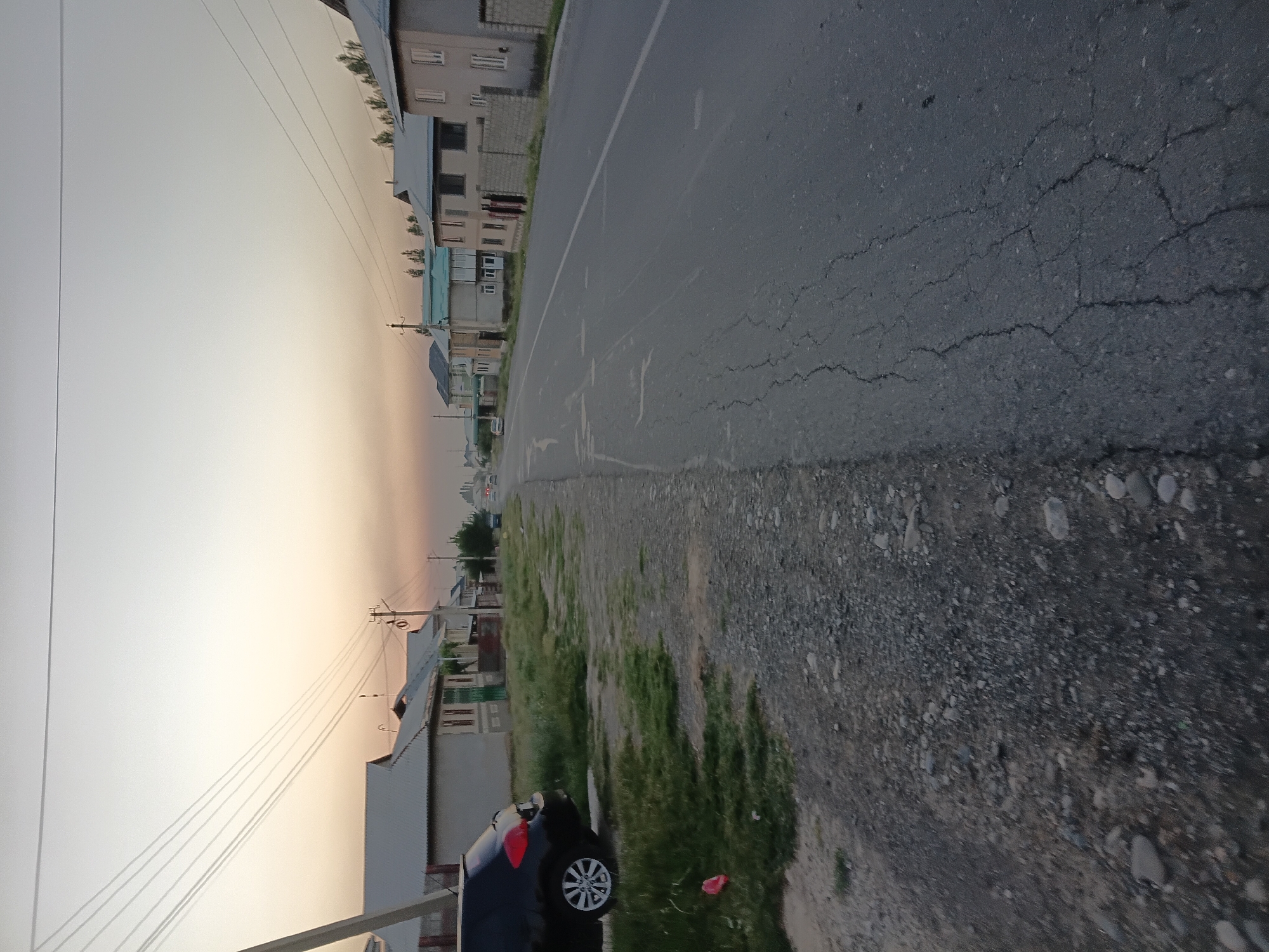 The image depicts a street scene in Uchkun, Kyrgyzstan, likely during the early morning or late evening due to the soft, diffused light and the warm hues in the sky. The perspective is from ground level, looking down a paved road that appears to be in moderate condition with visible cracks and some wear.
On the left side of the road, there is a grassy verge with scattered gravel and stones. A black car is parked on this verge, its rear visible, featuring a red taillight. Beyond the verge, a row of houses lines the street. These houses are of varied architectural styles, with some featuring light-colored facades and pitched roofs. One house has a green door.
The right side of the road is also lined with houses, some of which are taller and appear to be multi-story residential buildings. These also have light-colored walls and some have fencing. The sky above is a pale grey transitioning to a warm orange-yellow haze near the horizon, suggesting either sunrise or sunset. Power lines stretch across the upper portion of the image, supported by utility poles. In the distance, more houses and trees are visible, and there are a few other vehicles on the road, suggesting a populated area. The overall impression is a quiet, residential street in a provincial town.