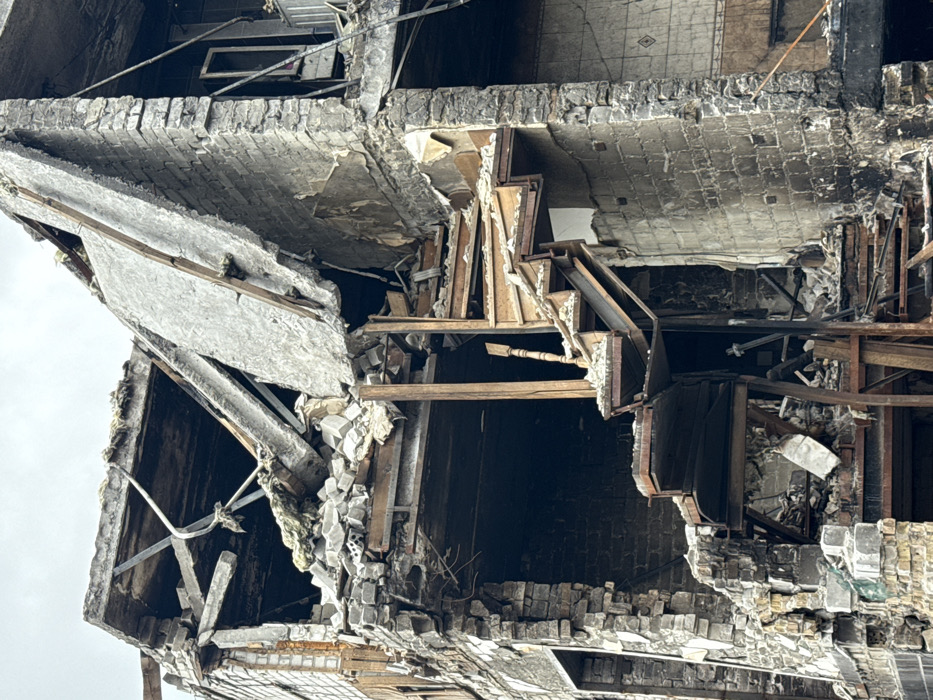 The image shows the aftermath of severe structural damage to a multi-story building. Debris is scattered, exposing layers of brick, concrete, and internal framing. Jagged edges and unstable-looking sections suggest a collapse or explosion. Interior features like remnants of stairs and flooring are visible within the wreckage, underscoring the destruction. The building's overall integrity is compromised, indicating a scene of significant devastation.