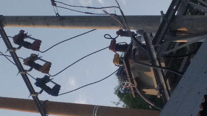 This image depicts electrical infrastructure on a clear day in Lamreung, Indonesia. Two utility poles are visible: a brown pole on the left and a gray concrete pole on the right. A metal crossarm is mounted between the poles, supporting three overhead primary cut-out fuses, colored black, yellow, and red from left to right. Electrical wires extend downwards from these fuses. Below the fuses, a large gray electrical transformer is mounted on a metal support structure positioned between the two poles. The transformer has various colored insulators (yellow and red are prominent) and multiple electrical cables connecting to and from it. Additional wires and cables are visible, running between the poles and the transformer. In the background, green tree foliage is present under a light blue sky with sparse white clouds. The corrugated dark gray roof of a building is visible in the lower left foreground. No individuals are present in the scene, and no active human actions or interactions are depicted.