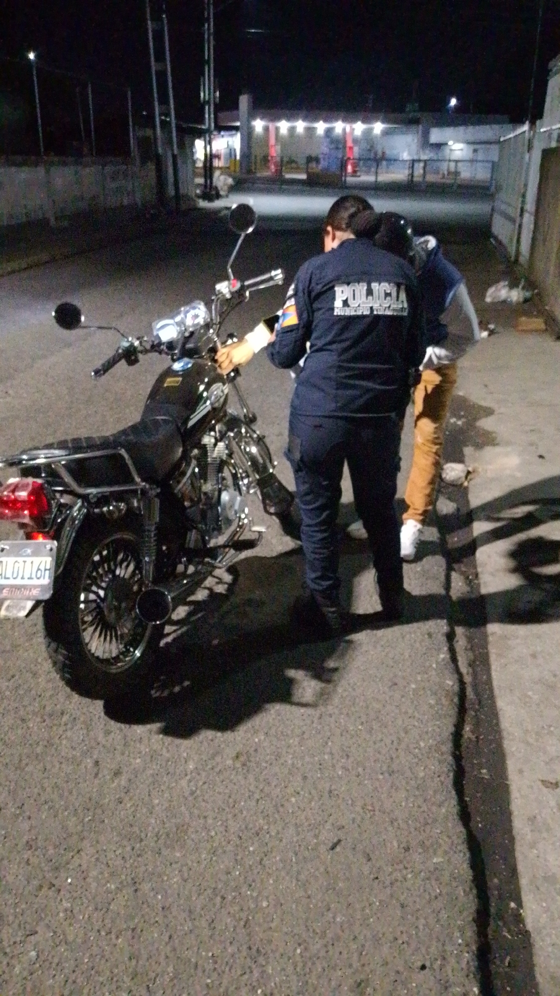 At night in what appears to be an alleyway or side street in Tinaquillo, Venezuela, a municipal police officer in full uniform (dark blue shirt with “POLICIA MUNICIPIO TINAQUILLO” written on the back) is inspecting a dark-colored motorcycle. The license plate is visible and reads “AL0116H”. The officer is wearing gloves. A person is standing next to the officer, but the details of this person's attire and face are obscured. Behind them, a building or structure is illuminated. A concrete wall bounds one side of the scene. The pavement is cracked and uneven.