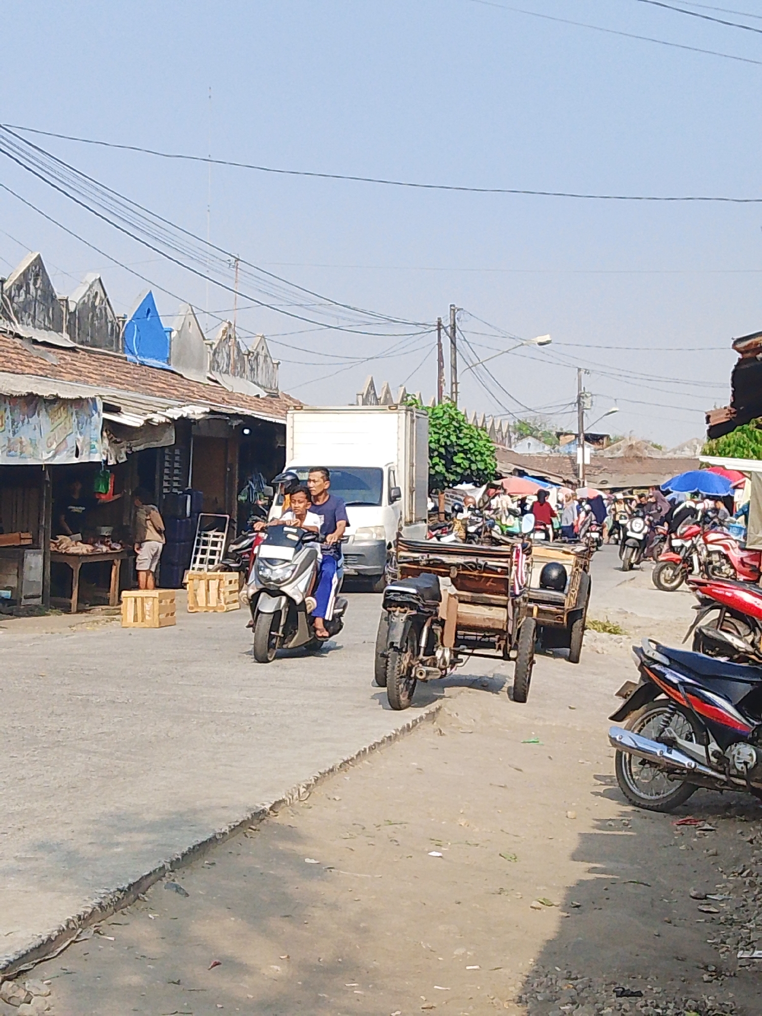 The image depicts a bustling street scene in Kalinyamatan, Indonesia, likely a market area given the presence of vendors, stalls, and various modes of transportation. A man and a boy are riding a scooter in the foreground, heading towards the right side of the frame. Behind them, a two-wheeled cart with a wooden carriage is being pulled. Further down the street, numerous motorcycles, some carrying passengers and others parked, are visible. Several stalls with awnings are set up along the roadside, with people engaged in transactions. A white truck is parked in the background. The environment is characterized by a paved road, sandy shoulders, and a clear sky. Overhead, a network of electrical wires crisscrosses the sky. The architecture of the buildings suggests a warm climate, with tiled roofs and open-fronted stalls. The overall atmosphere is one of daily commerce and movement within a community.
