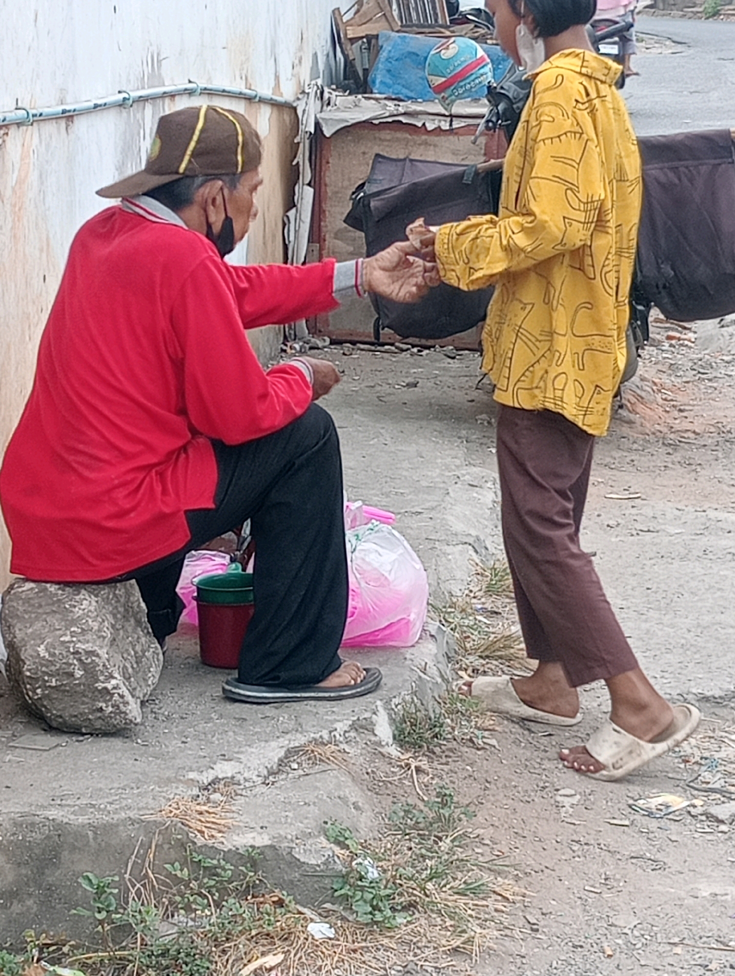 An older person wearing a red long-sleeved shirt, black pants, and a brown baseball cap sits on a large rock by a concrete curb in Bandar Lampung, Indonesia. The person is wearing a black face mask and appears to be handing something to a younger person. The younger person, wearing a yellow shirt with a black line pattern and brown pants, stands facing the older person and is wearing white sandals. Next to the seated person is a red bucket with a green lid and a clear plastic bag containing pink items. The ground is dirt and gravel, with some dry grass and small green plants growing near the curb. In the background, there are structures made of wood and metal, with tarps and bags, suggesting an informal market or roadside setup. A helmet with a cartoon character design is visible in the background, along with a motorcycle. The wall behind the seated person is light-colored with blue pipes running along it.