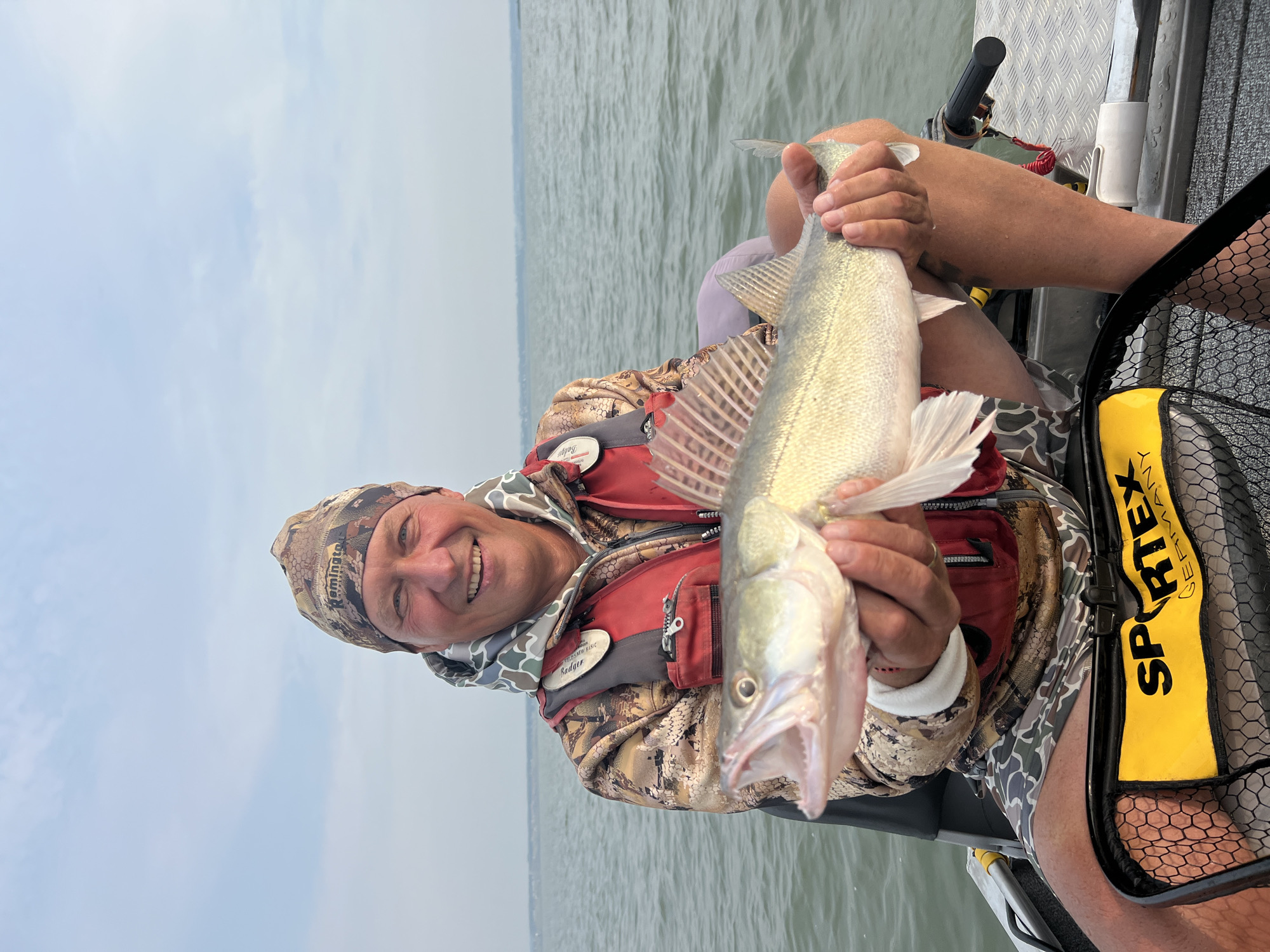 A middle-aged Caucasian man, wearing a camouflage beanie, a red life vest over a camouflage hoodie, and camouflage shorts, sits in a boat on a body of water. He is holding a freshly caught fish, a zander, with its mouth open, by its gills and tail. The man is smiling and looking at the camera. The background shows a hazy sky and a distant shoreline. A fishing net with a yellow bag labeled "SPORTEX GERMANY" is visible in the foreground. The scene appears to be a recreational fishing outing.