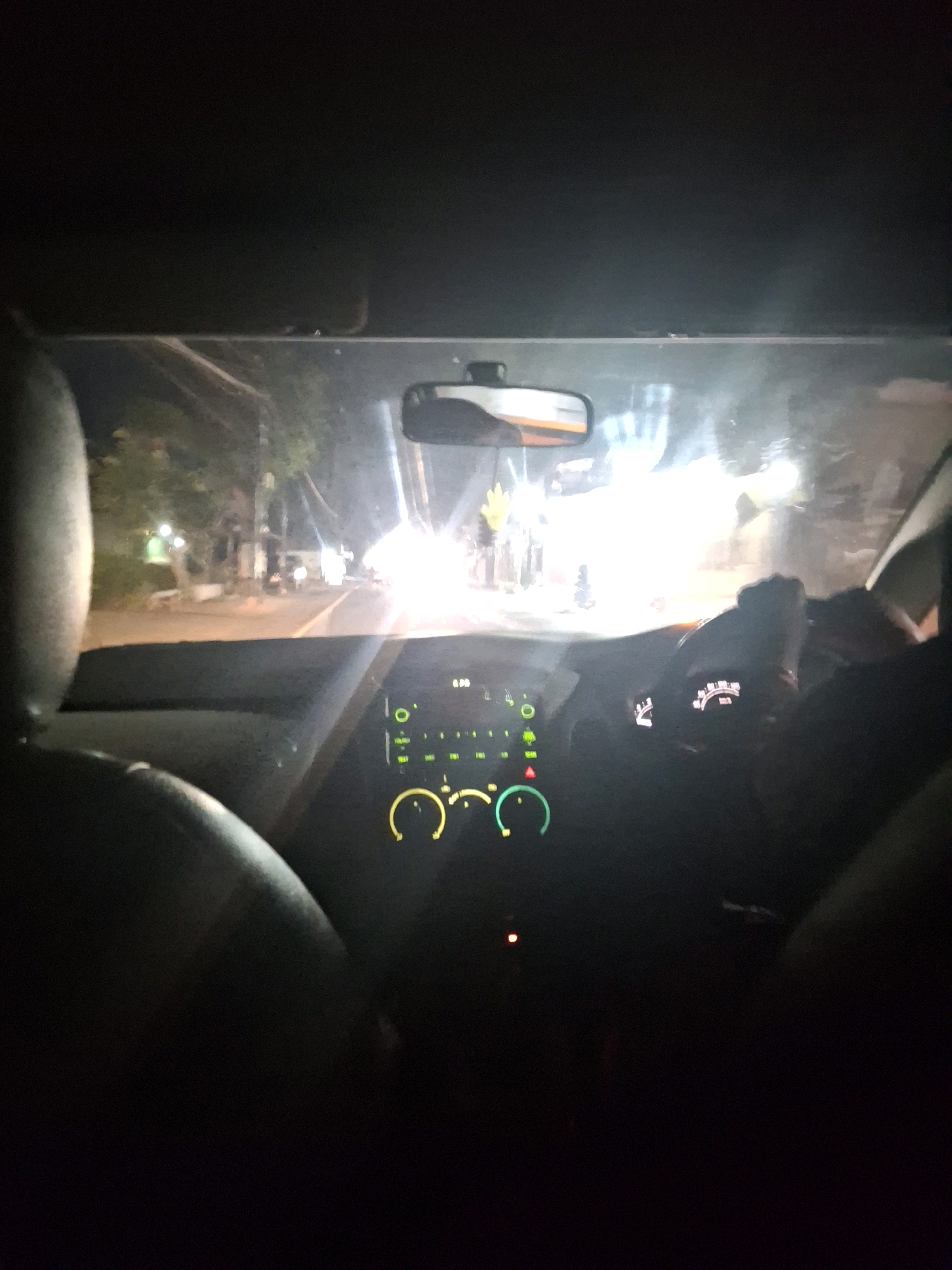 The image is a night-time, interior view from the passenger seat of a car, looking forward through the windshield. The car is on a street in Lumajang, Indonesia, as indicated by the context of the city. Outside the car, the street is illuminated by streetlights and the headlights of other vehicles, creating a bright, somewhat hazy glow. There are trees and buildings visible along the side of the road. The car's dashboard is dimly lit, with illuminated gauges and indicators. The steering wheel is partially visible, and a driver's hand rests on it. The overall impression is of a night drive through a moderately lit urban or suburban area.