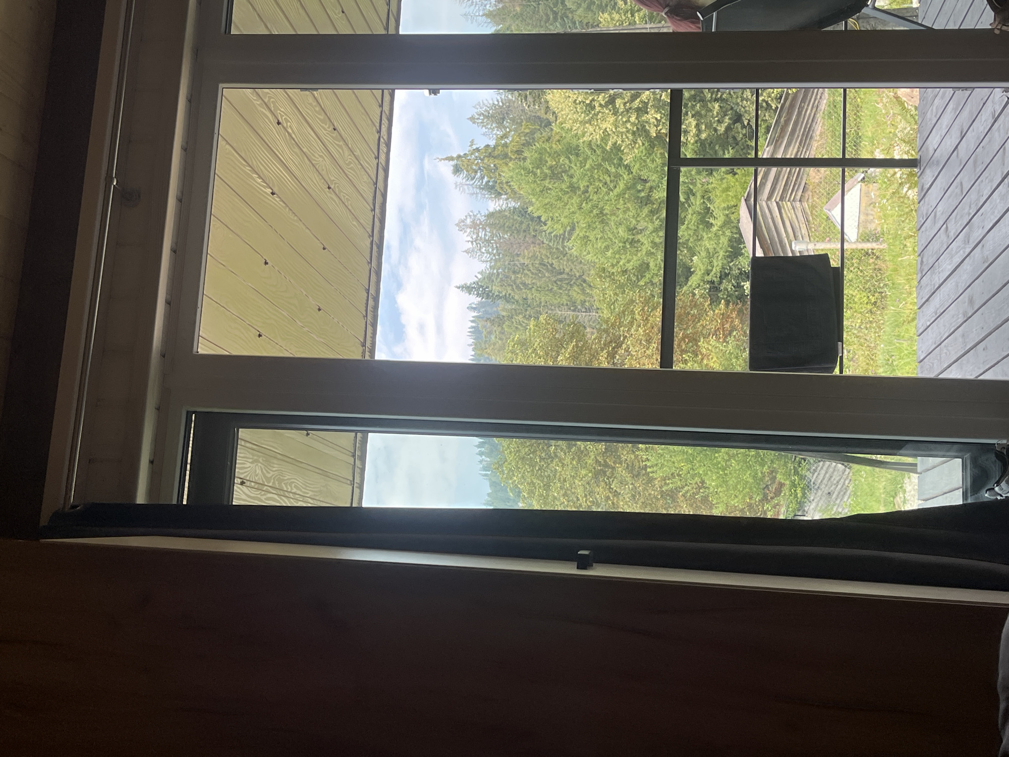 The image shows a view through a large window or sliding glass door of a room, looking out onto a wooded hillside. The room has wooden walls and a dark curtain partially drawn to the left. Outside, there is a wooden deck with a railing. Beyond the deck, a dense forest of green trees covers the hillside, with some of the trees appearing to be coniferous. In the mid-ground, there are a couple of rustic wooden buildings visible among the trees, one with a distinct stepped roof. The sky above is partly cloudy. A dark towel is hanging on the railing. The overall scene suggests a natural, possibly remote, setting. It is likely located in or near Mykulychyn, Ukraine, given the mention of the city.