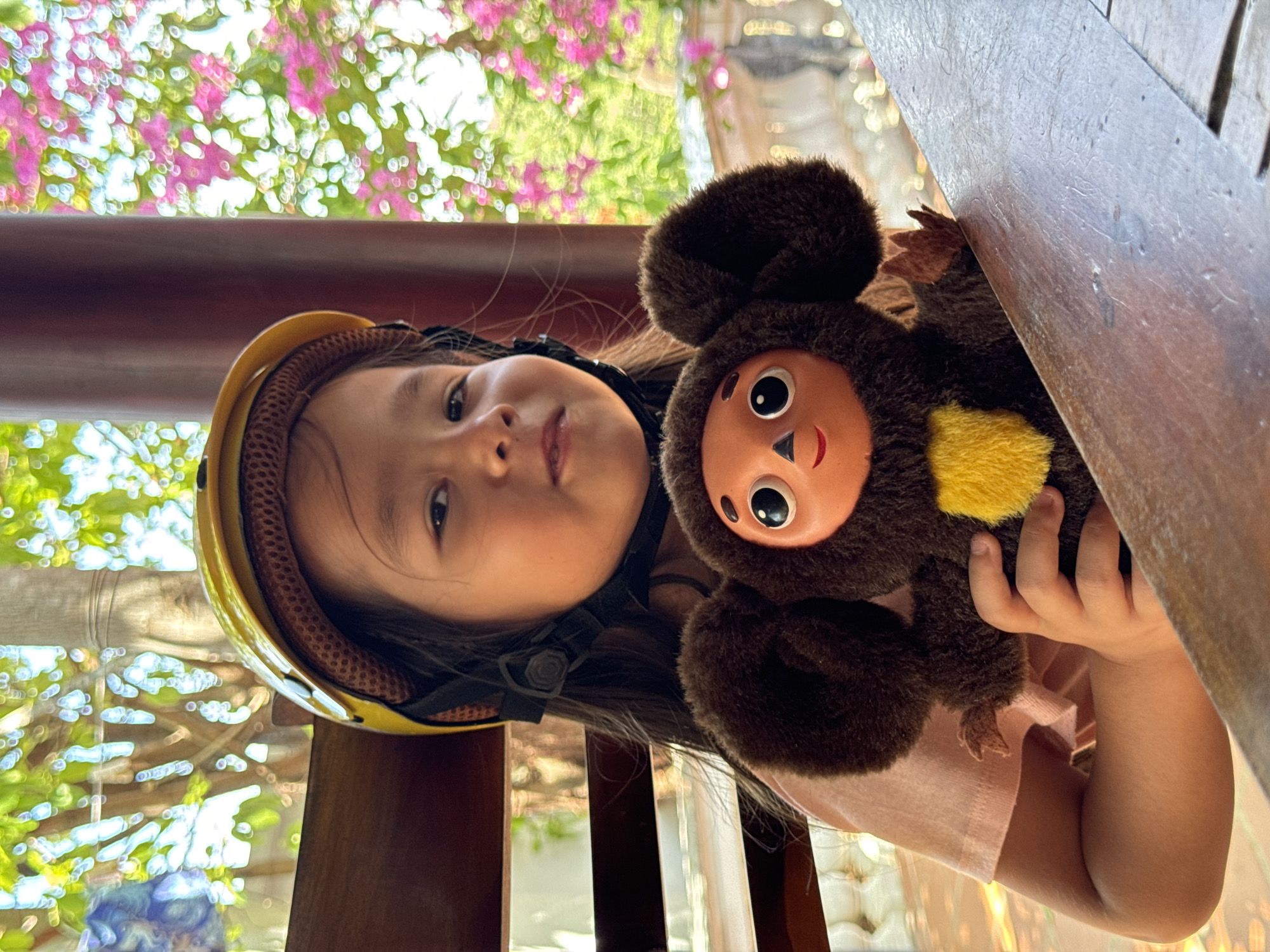 A young Asian girl wearing a yellow helmet with a black strap poses indoors with a Cheburashka plush toy. The girl holds the toy close to her chest. Visible through the background are flowers and a blurred cityscape that suggests this indoor location might be on a veranda or sun porch. The city is identified as Mũi Né, Vietnam.